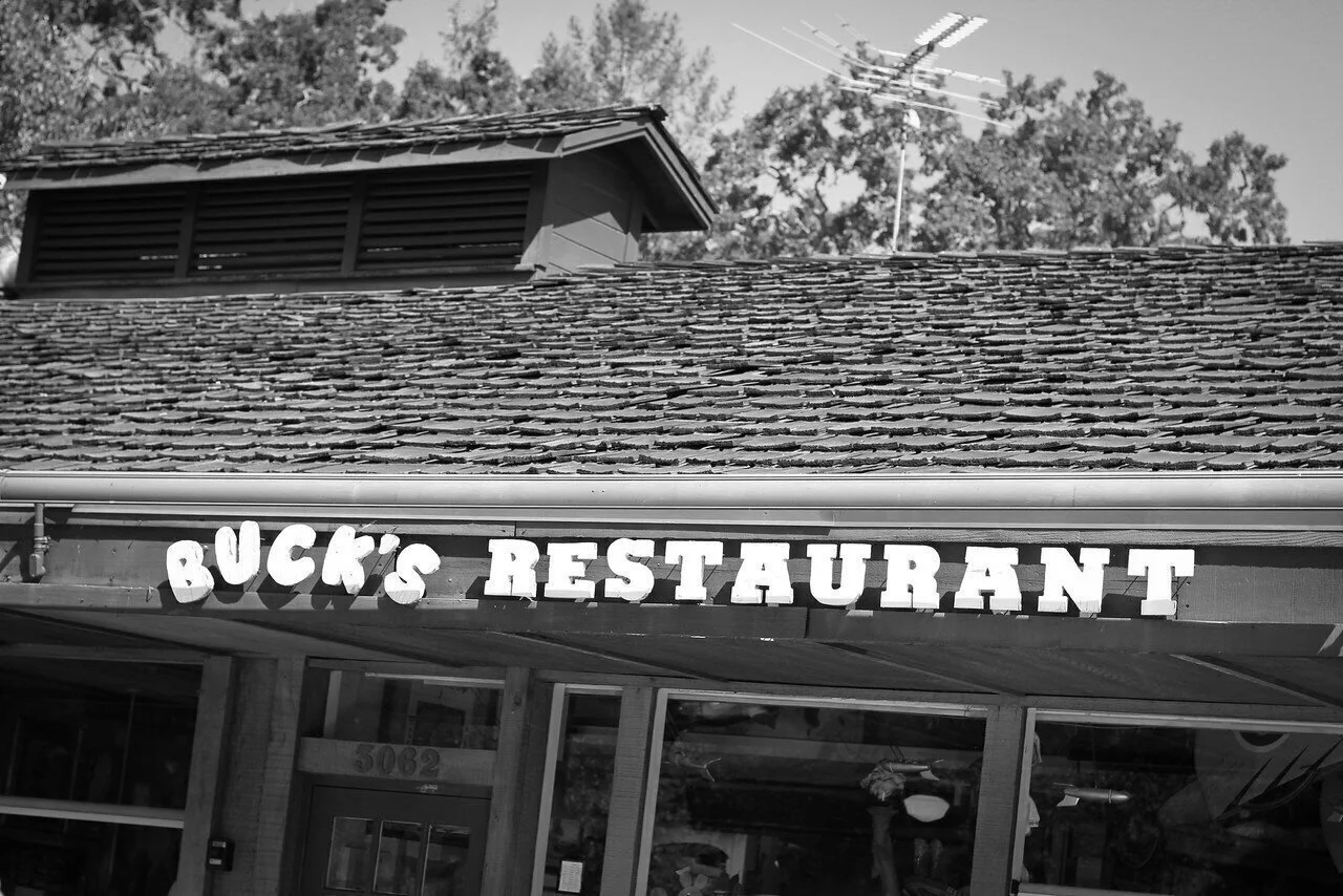 Black and white photo of a restaurant with a sign that reads "Buck's Restaurant" above the entrance, with trees and rooftops in the background.