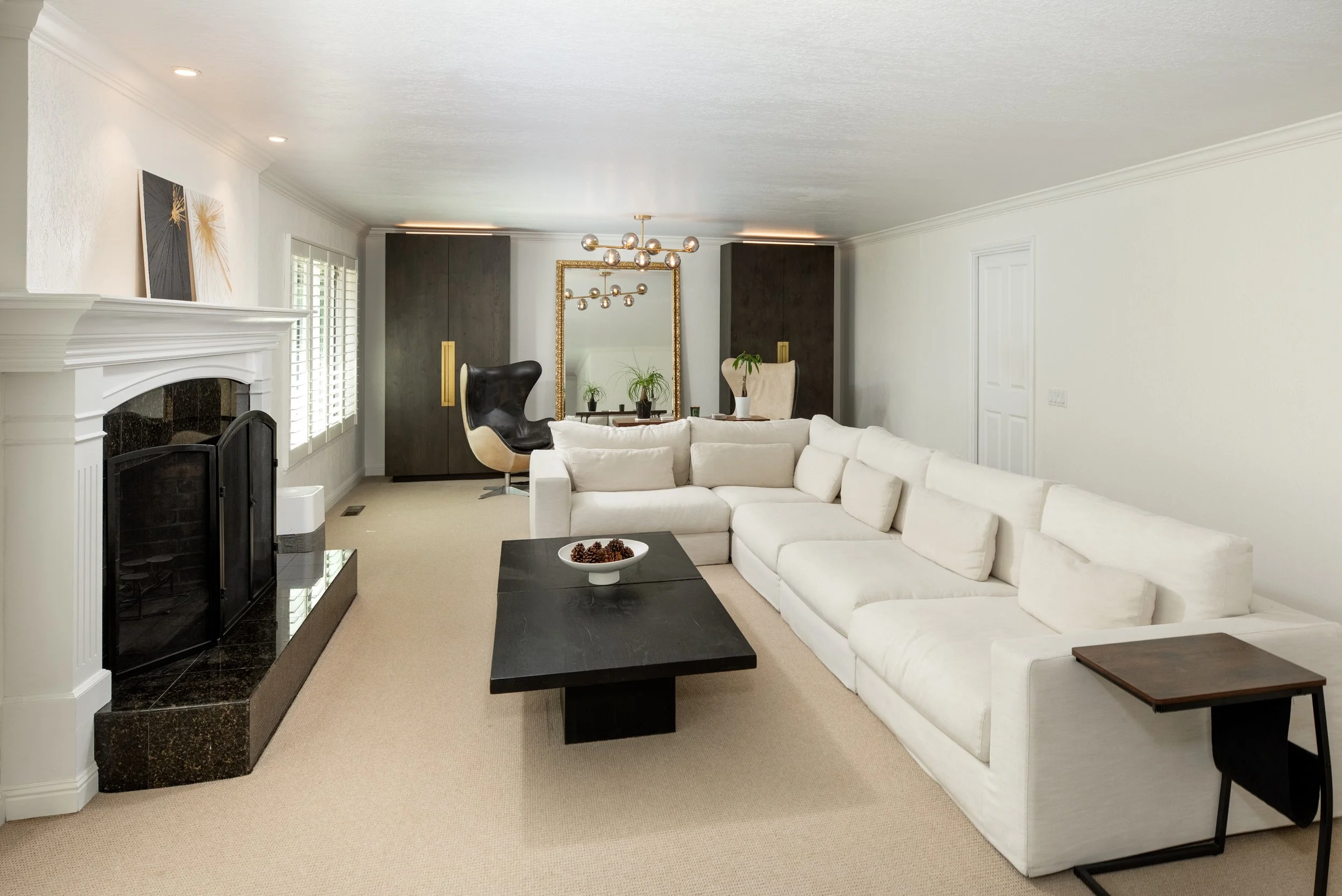 Living room with white sofa, black coffee table with decorative bowl, fireplace, large mirror with gold frame, modern chandelier, two black and beige chairs, and abstract paintings on the wall.