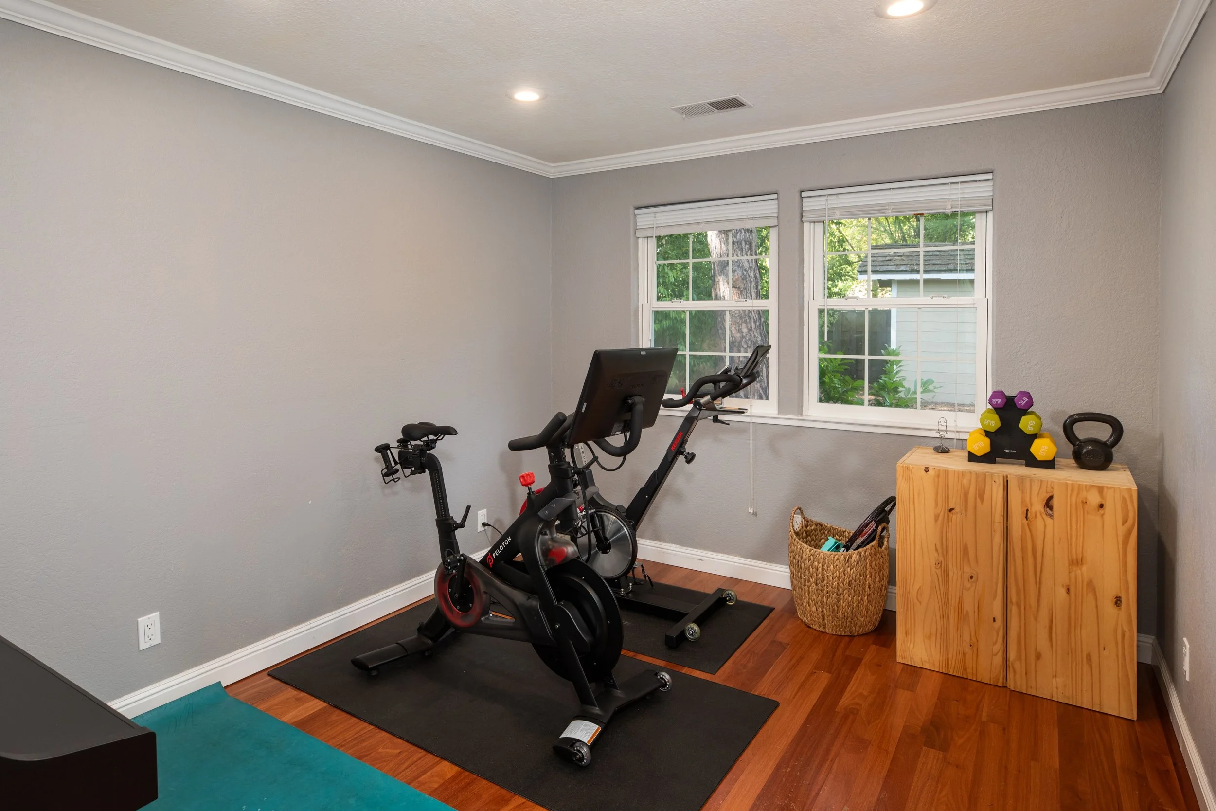Home gym with a Peloton exercise bike on a black mat, workout equipment including a kettlebell, a basket, and a wooden table with colorful dumbbells, two windows with blinds, gray walls, hardwood floor, and ceiling lights.