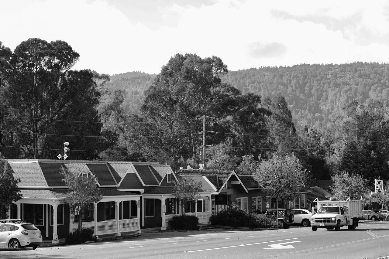 A street view in a small town with houses, parked cars, trees, power lines, and a mountainous background in black and white.