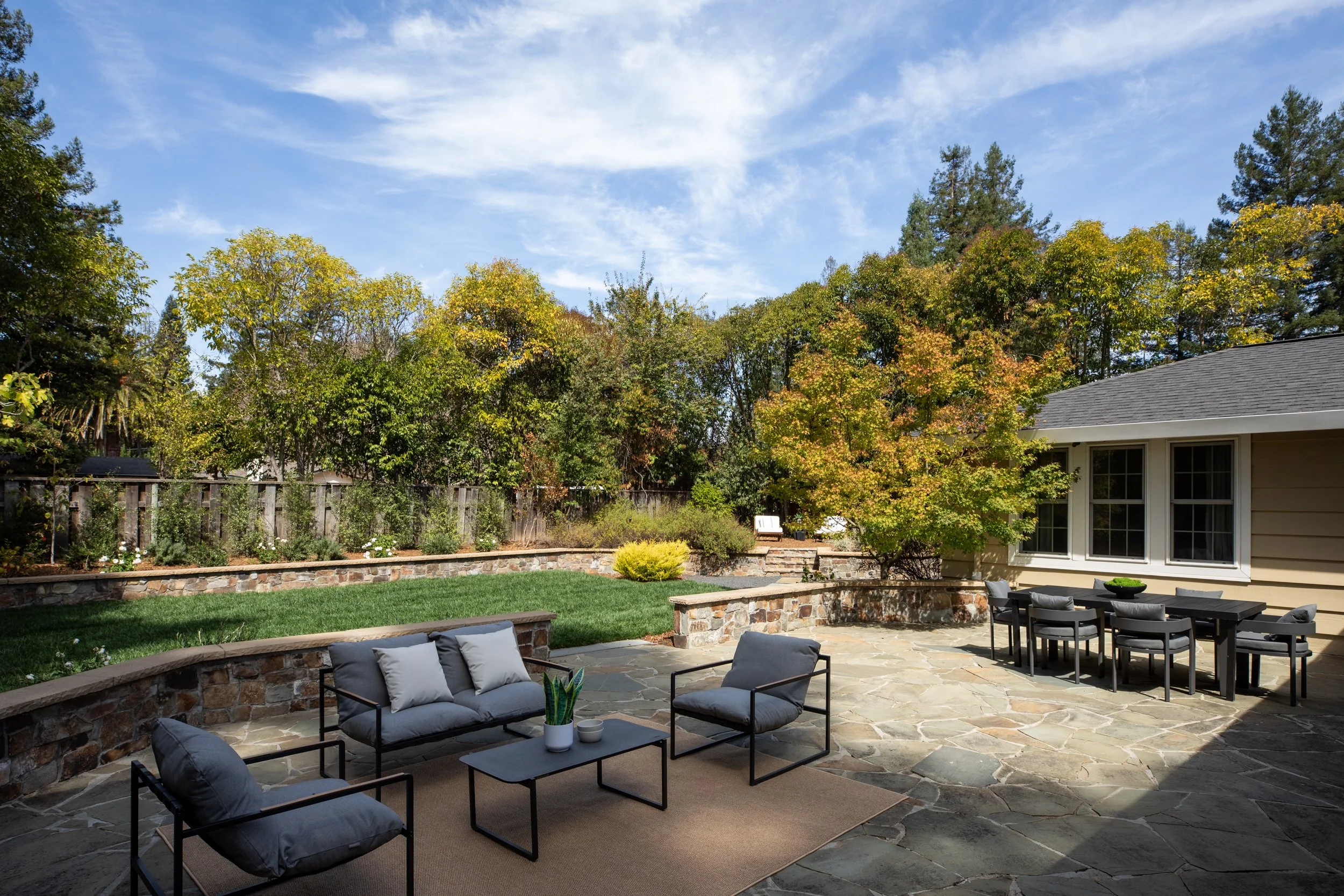 A backyard patio with outdoor seating, a built-in stone fireplace, lush green lawn, and various trees with some showing fall foliage. A house is partially visible on the right side.