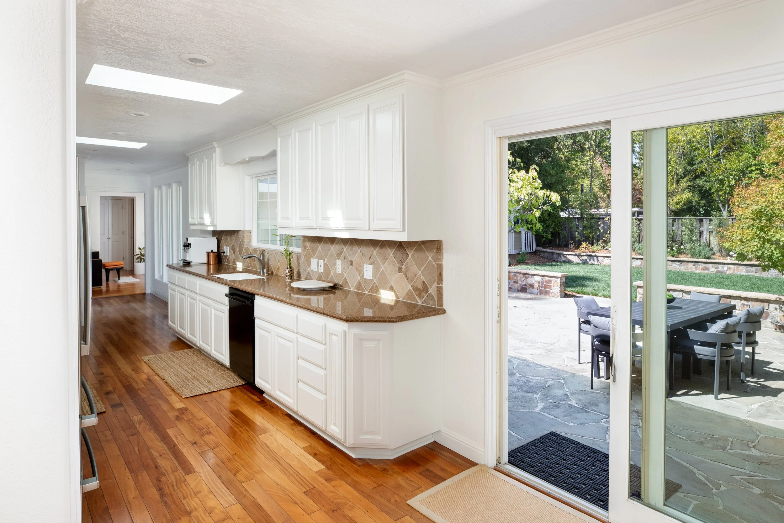 A view of a bright kitchen with white cabinets, a brown tile backsplash, and a granite countertop. There is a sliding glass door leading to an outdoor patio with a table and chairs, and a backyard with trees.