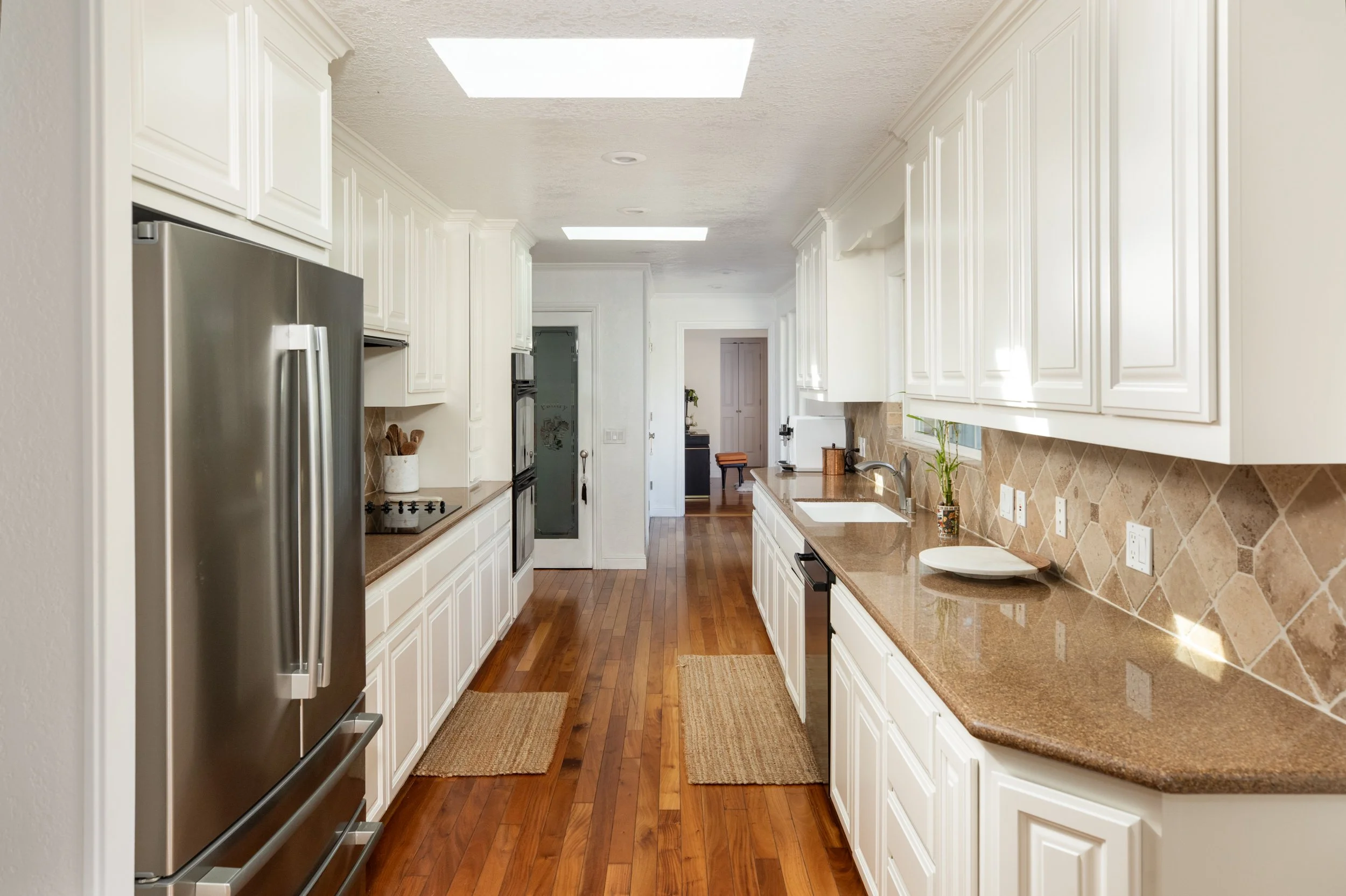 A long kitchen with white cabinets, brown countertops, wood flooring, and a stainless steel refrigerator on the left. There are two rugs on the floor, and a windowed upper ceiling light.