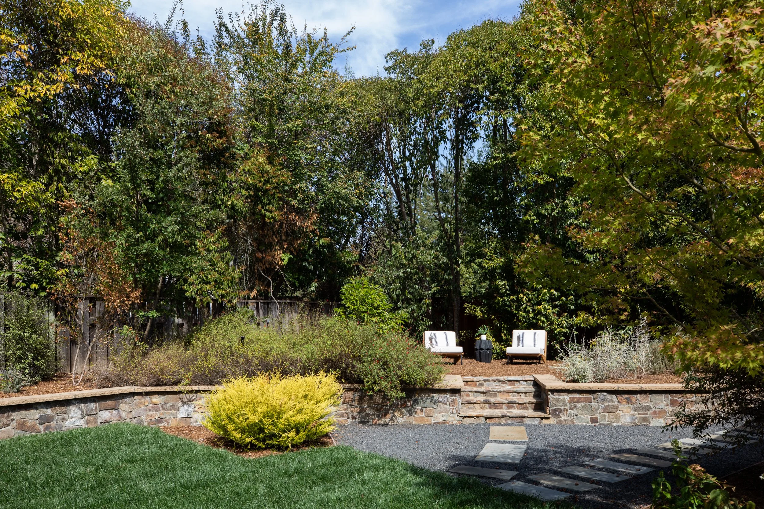 A backyard garden with lush green grass, yellow and green bushes, and tall trees with green and reddish leaves. Two white chairs are placed on the patio, and a stone retaining wall lines the garden. Blue sky with some clouds is visible in the backgro
