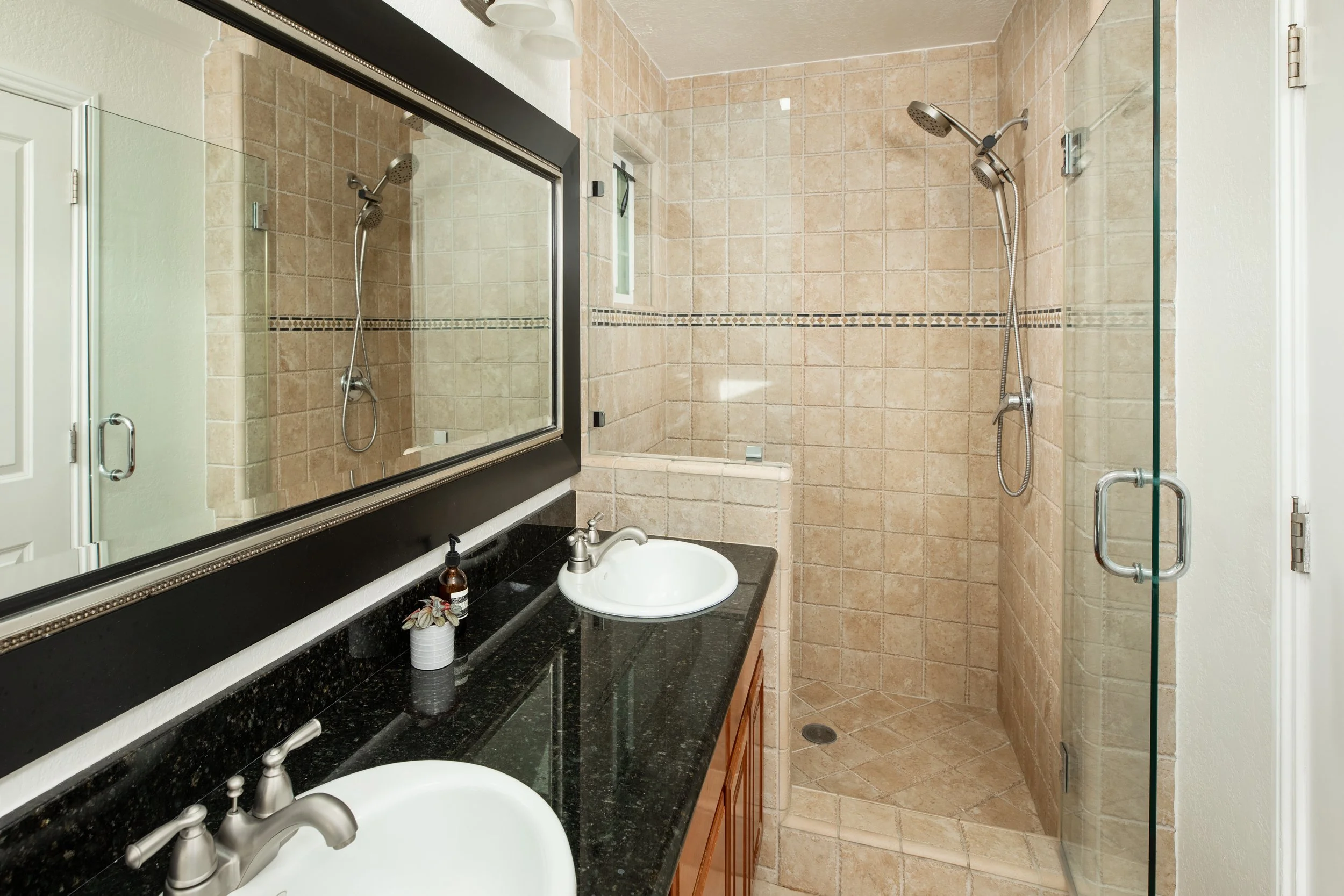A bathroom with beige tiled walls and a glass shower enclosure featuring dual showerheads. There is a black granite countertop with two white sinks and silver fixtures, a large mirror with a black frame above the countertop, and a small window in the