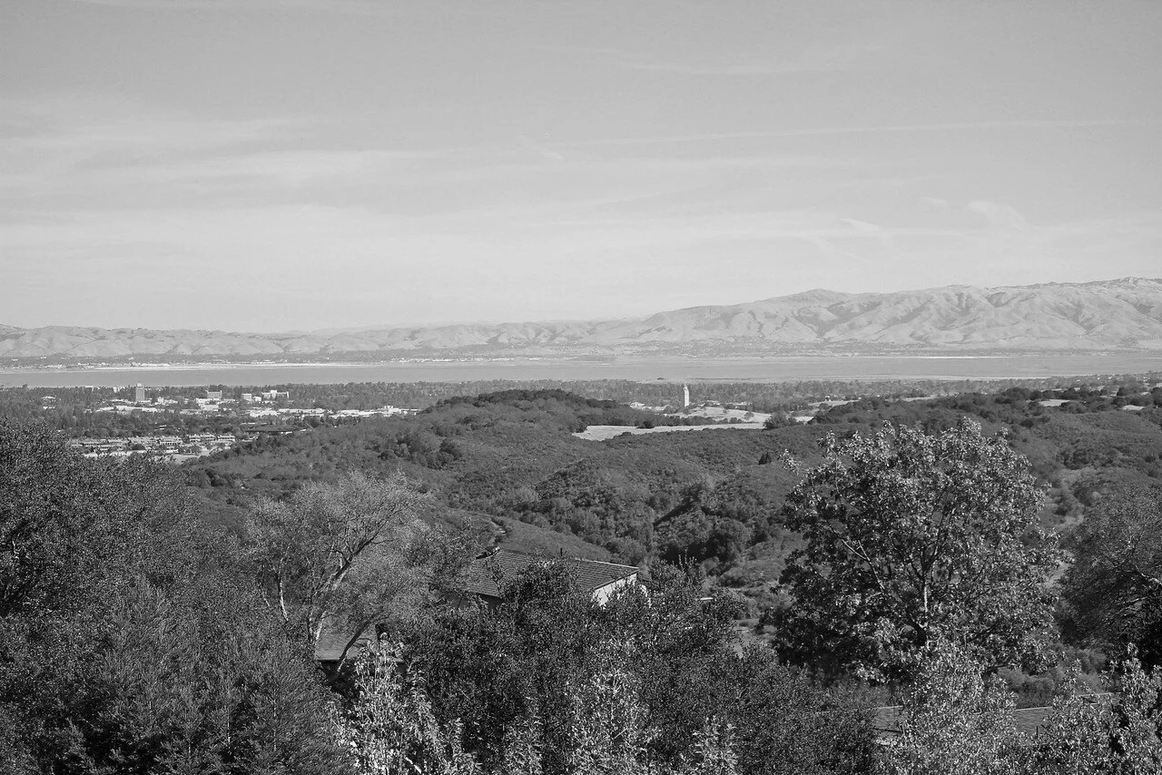 A black and white landscape photo showing rolling hills with trees in the foreground, a city or town in the middle ground, and mountains in the background under a partly cloudy sky.