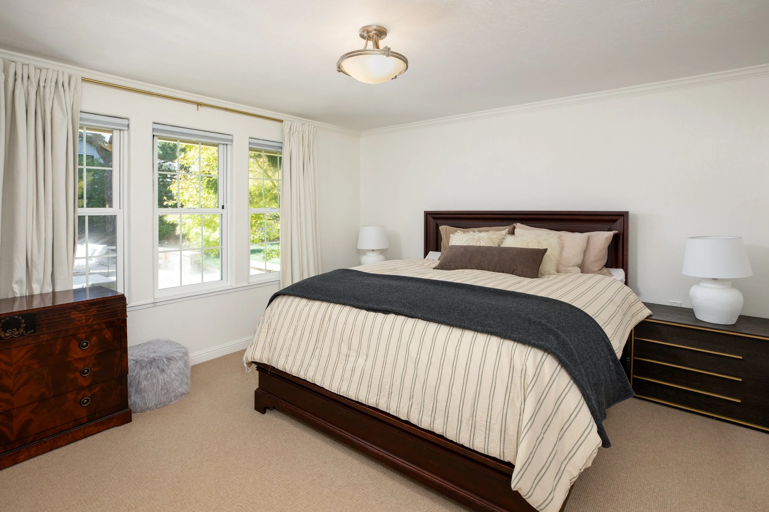 A bedroom with a large bed featuring a wooden headboard, striped bedding, and multiple pillows. There are two nightstands with lamps on either side of the bed. The room has three windows with white curtains, and outside greenery is visible. An elegan
