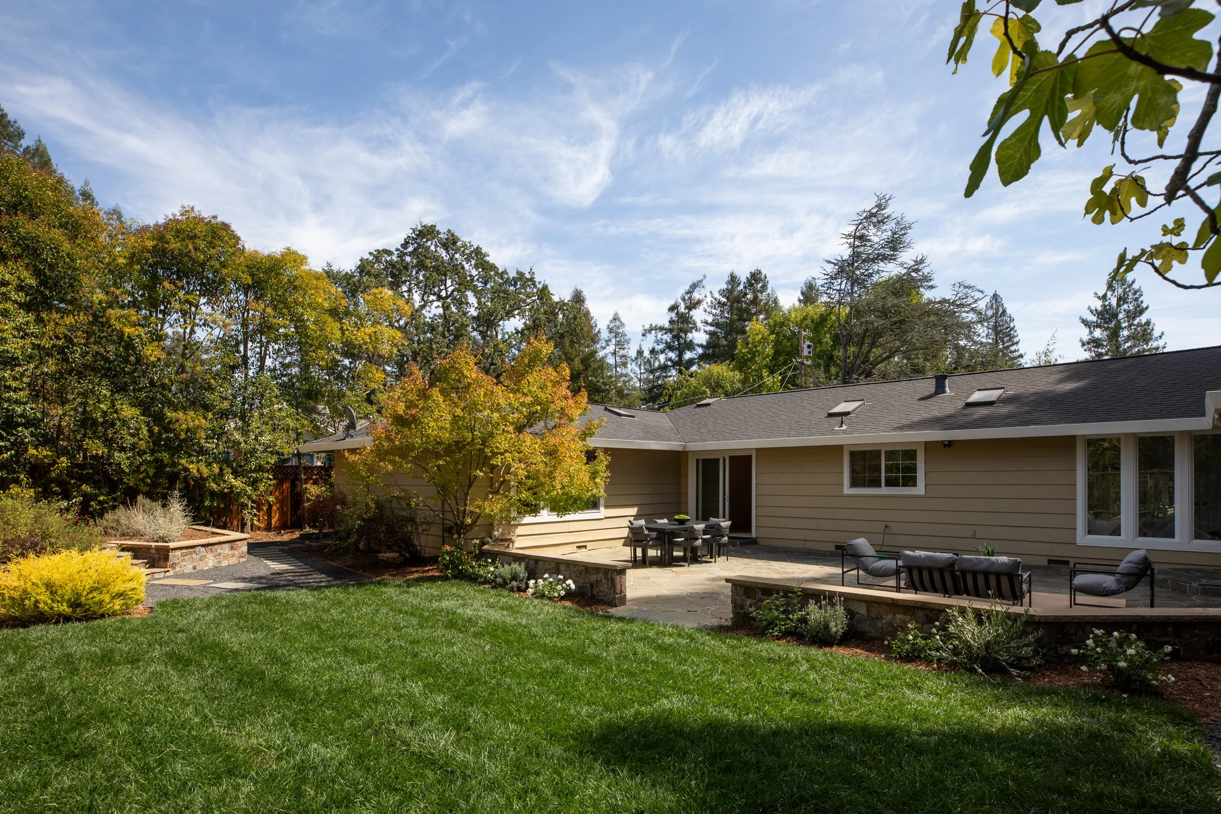 Backyard patio area with outdoor furniture, surrounded by green grass, shrubs, and trees, with a house in the background under a partly cloudy sky.