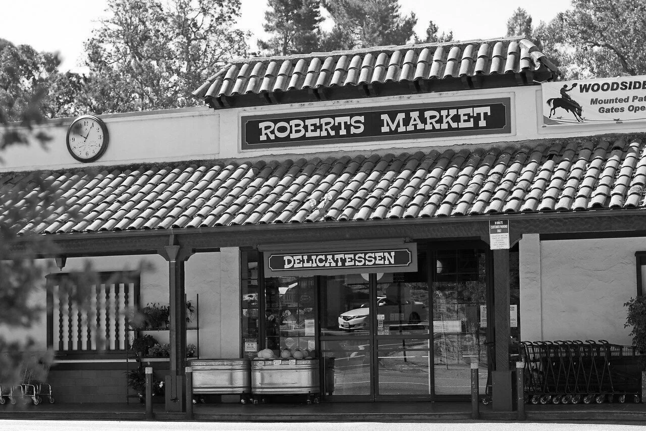 Front view of Roberts Market, a small deli with a tiled roof, a clock showing 3:05, shopping carts outside, and a sign indicating a delicatessen.