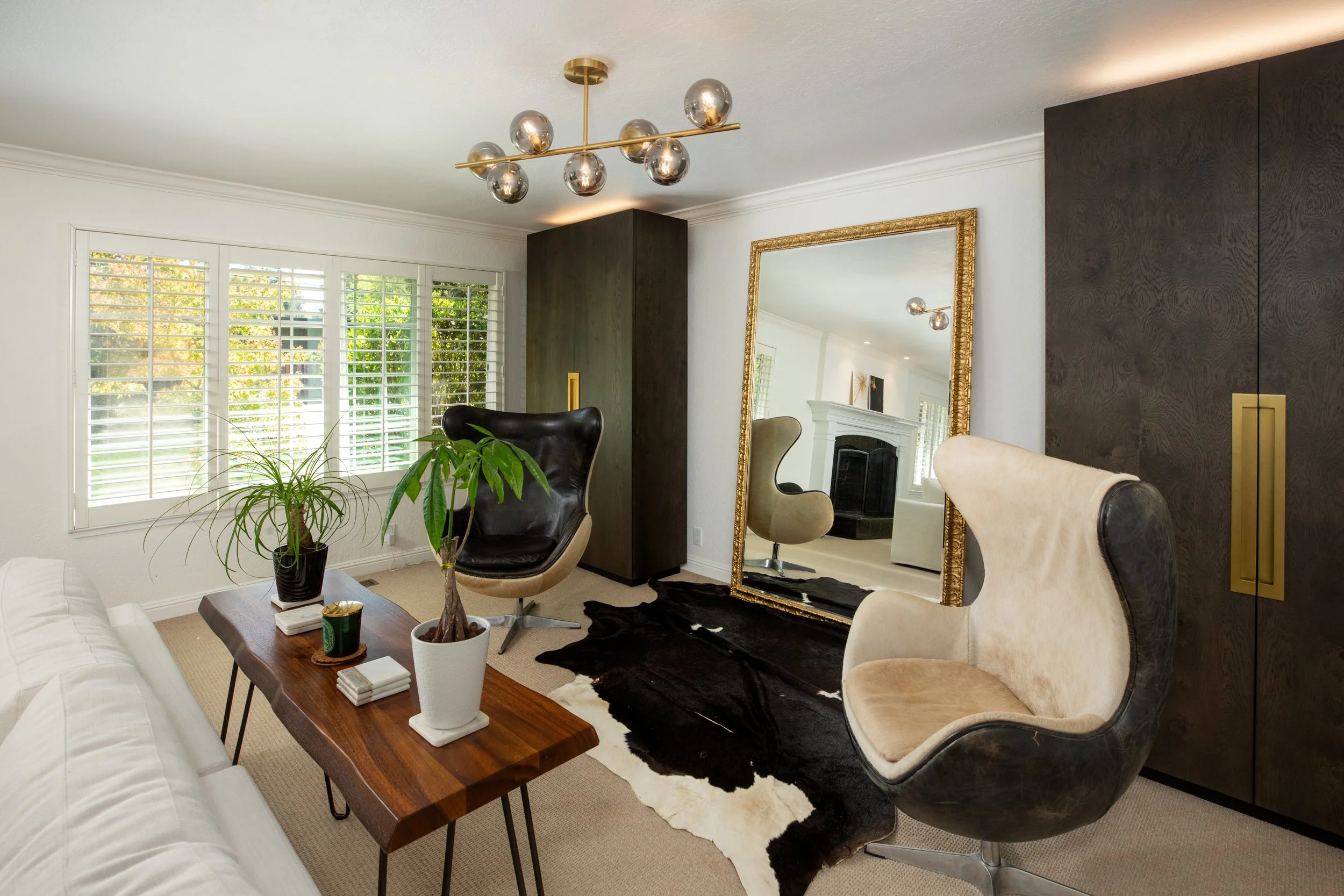 Living room with white walls, large window with shutters, modern gold chandelier, black and beige lounge chairs, large gold-framed mirror, dark wood cabinet, houseplants, and various furniture pieces.