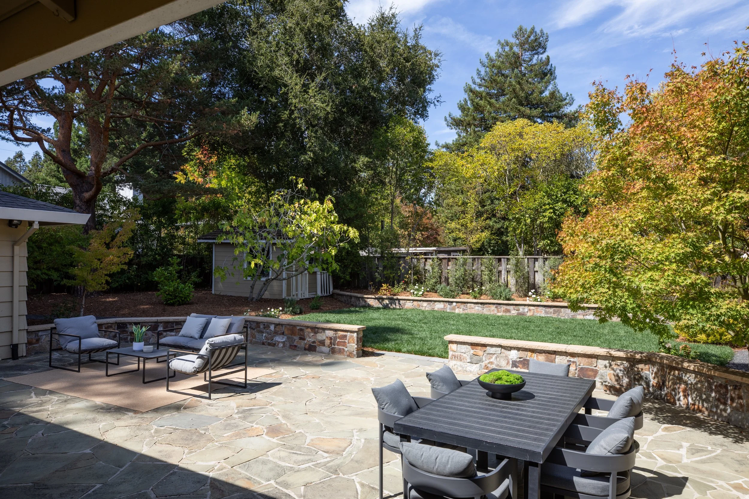 Backyard patio with outdoor seating, stone paved ground, lush green trees, and a clear blue sky.