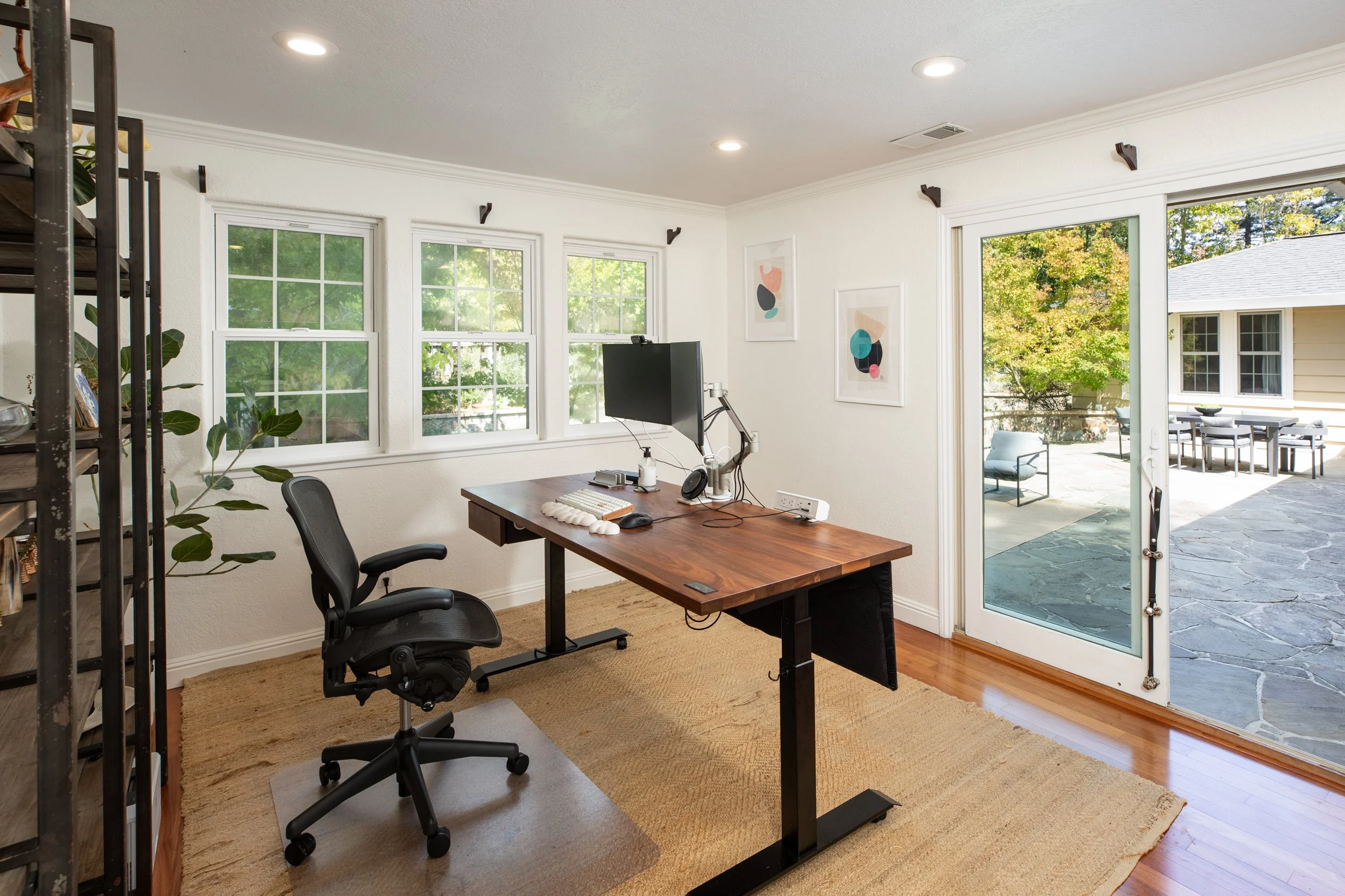 Home office with white walls, large windows, and a glass door leading to an outdoor patio. Contains a wooden desk with a computer monitor, keyboard, mouse, headphones, and a desk chair. There is a black metal shelving unit on the left side and framed