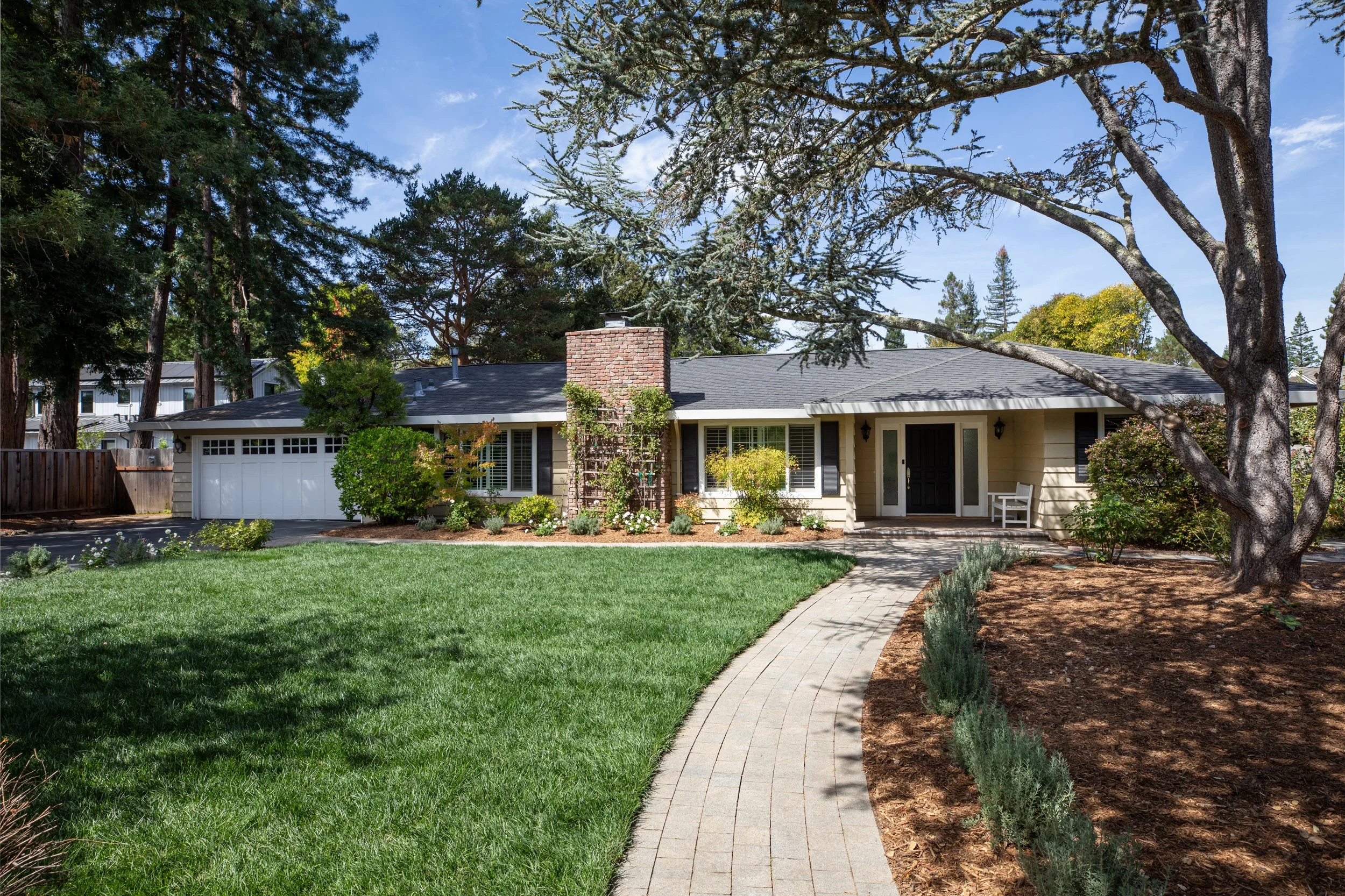 A single-story house with a dark gray roof, brick chimney, and white siding, situated in a lush green yard with a curved walkway, surrounded by trees and shrubs, under a blue sky.