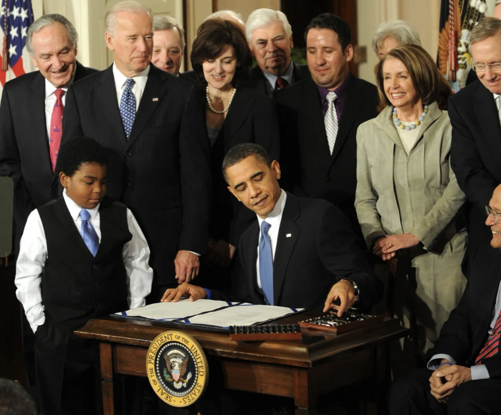 President Barack Obama signing a document at a desk with the presidential seal, surrounded by a diverse group of adults, some smiling and watching closely, in a formal setting.