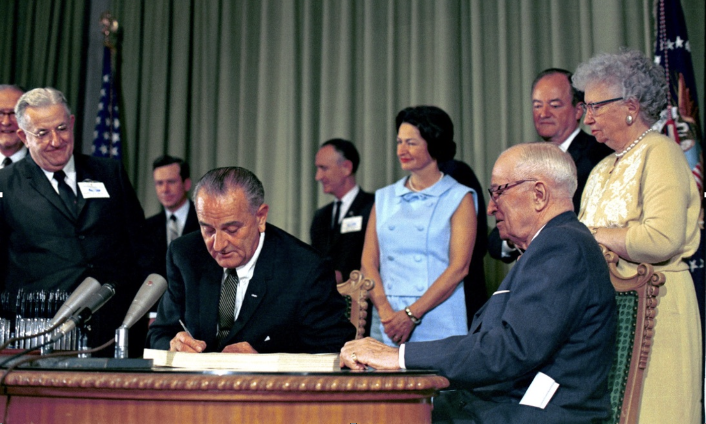 President Lyndon B. Johnson signing a document at a table, surrounded by officials and observers, during a formal signing ceremony in a room with green curtains and American flags in the background.