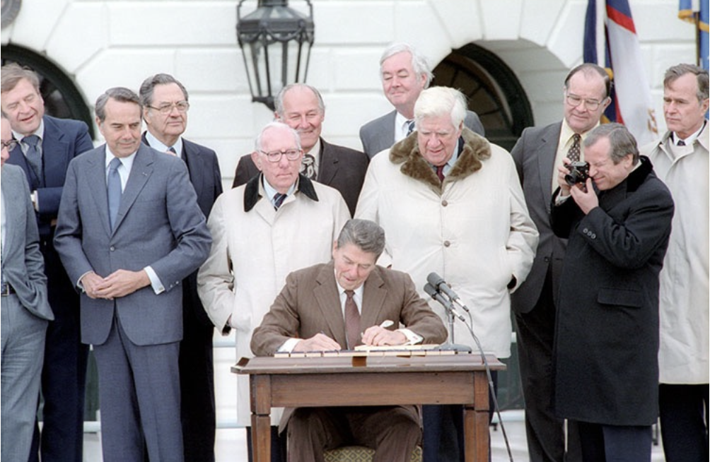 A man seated at a desk signing a document, surrounded by several men in formal and semi-formal attire, outdoors in front of the White House with a flag and lantern visible.