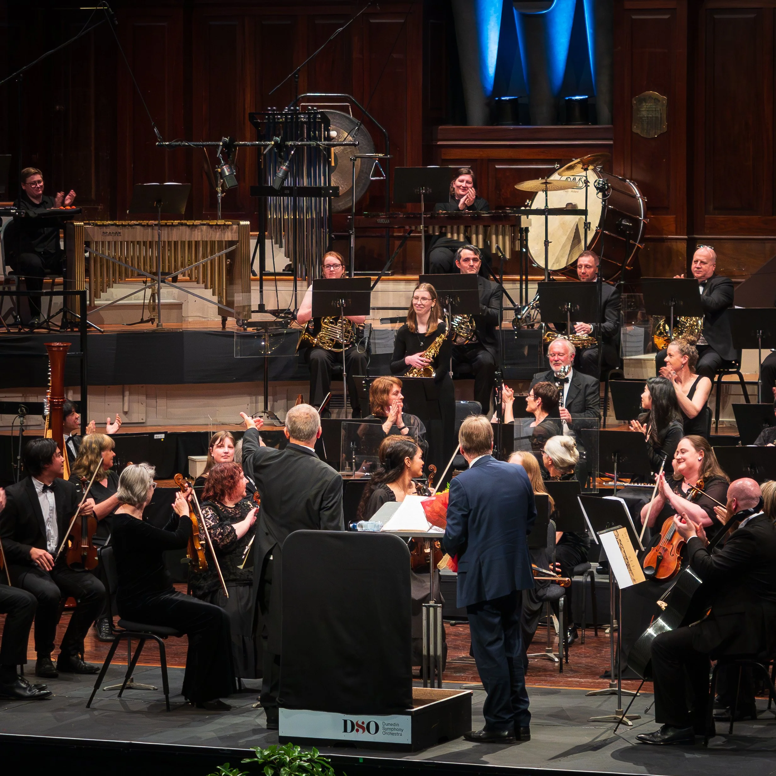 Orchestra members on stage after a performance at the Dunedin Symphony Orchestra, clapping and smiling, with musical instruments including drums, cymbals, and a vibraphone visible.