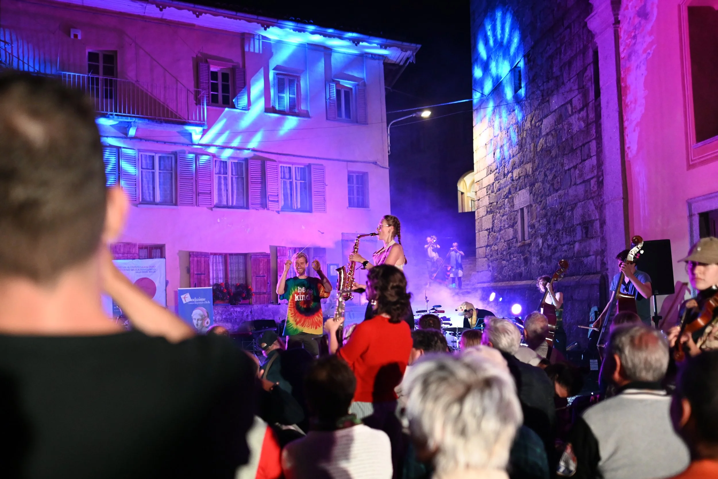 A nighttime outdoor concert with a band performing on a stage illuminated by colorful purple and blue lights. Audience members are visible in the foreground, some wearing bright clothing, and a building with windows and shutters is in the background.