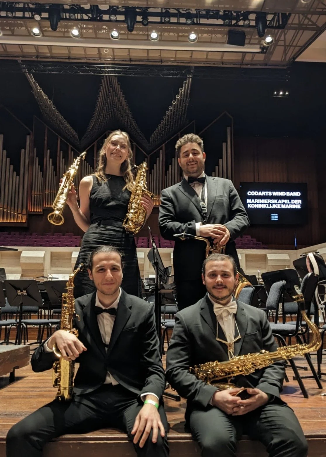 A group of five musicians dressed in formal black attire with white shirts, some with bow ties, pose with their musical instruments in a concert hall. The setting includes an organ in the background and chairs for the orchestra. There is a screen displaying text about the wind band.