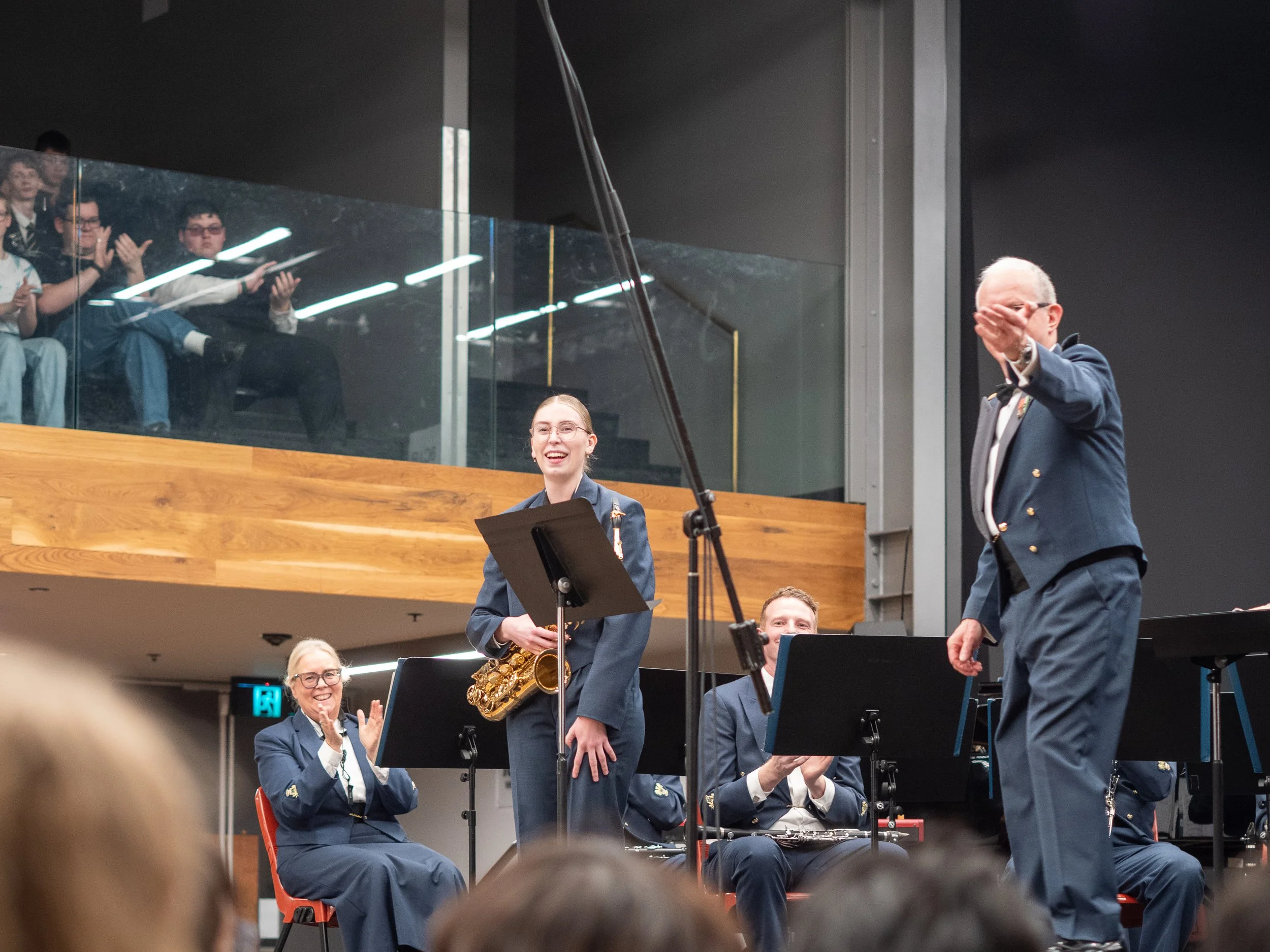 A group of musicians in uniform, some standing and some sitting, are performing on stage at a concert hall. The musicians are clapping and smiling, while one person is playing a saxophone. The audience is visible in the foreground.