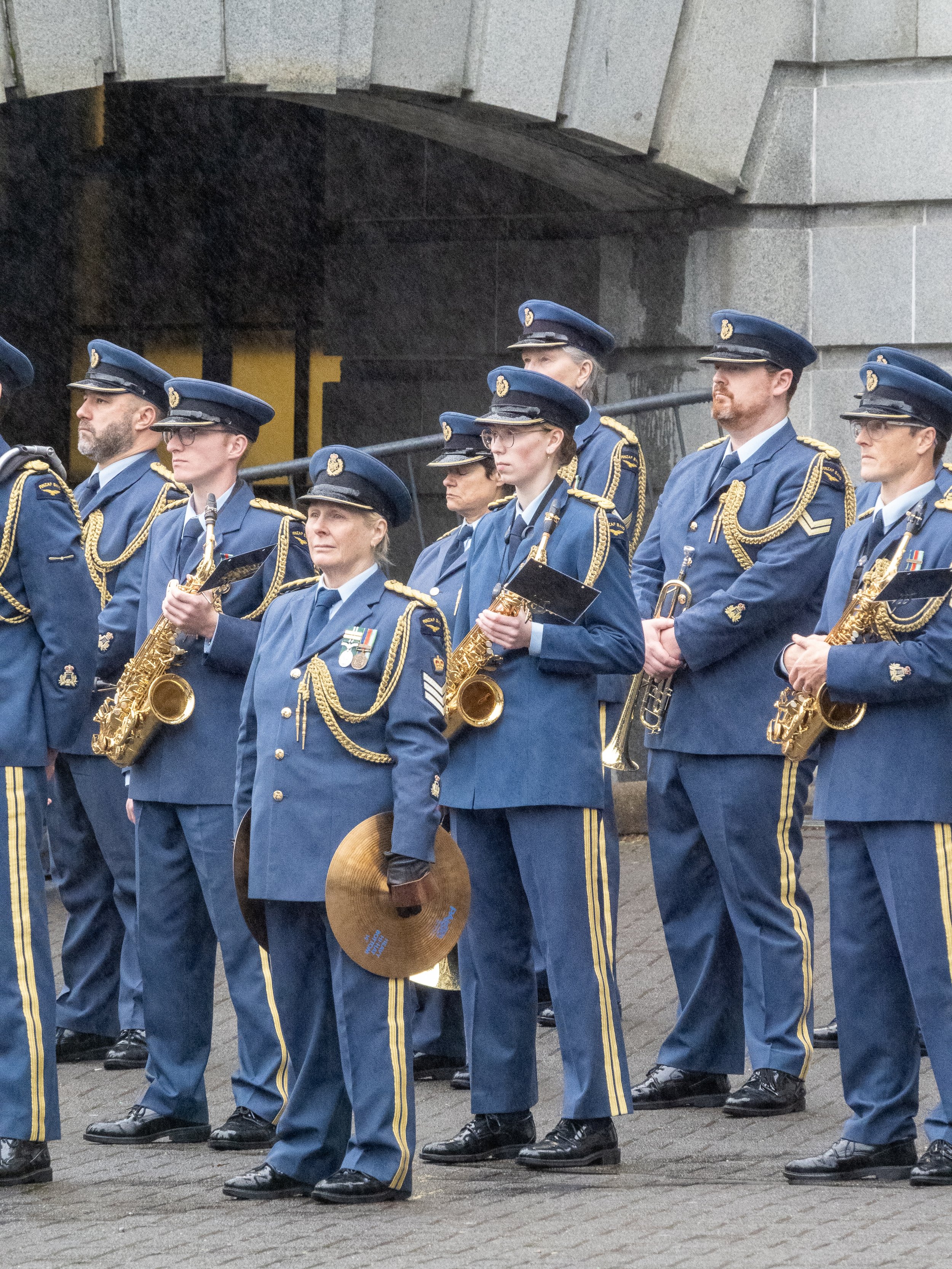 Group of uniformed military musicians standing in formation, holding musical instruments, in front of a stone wall.