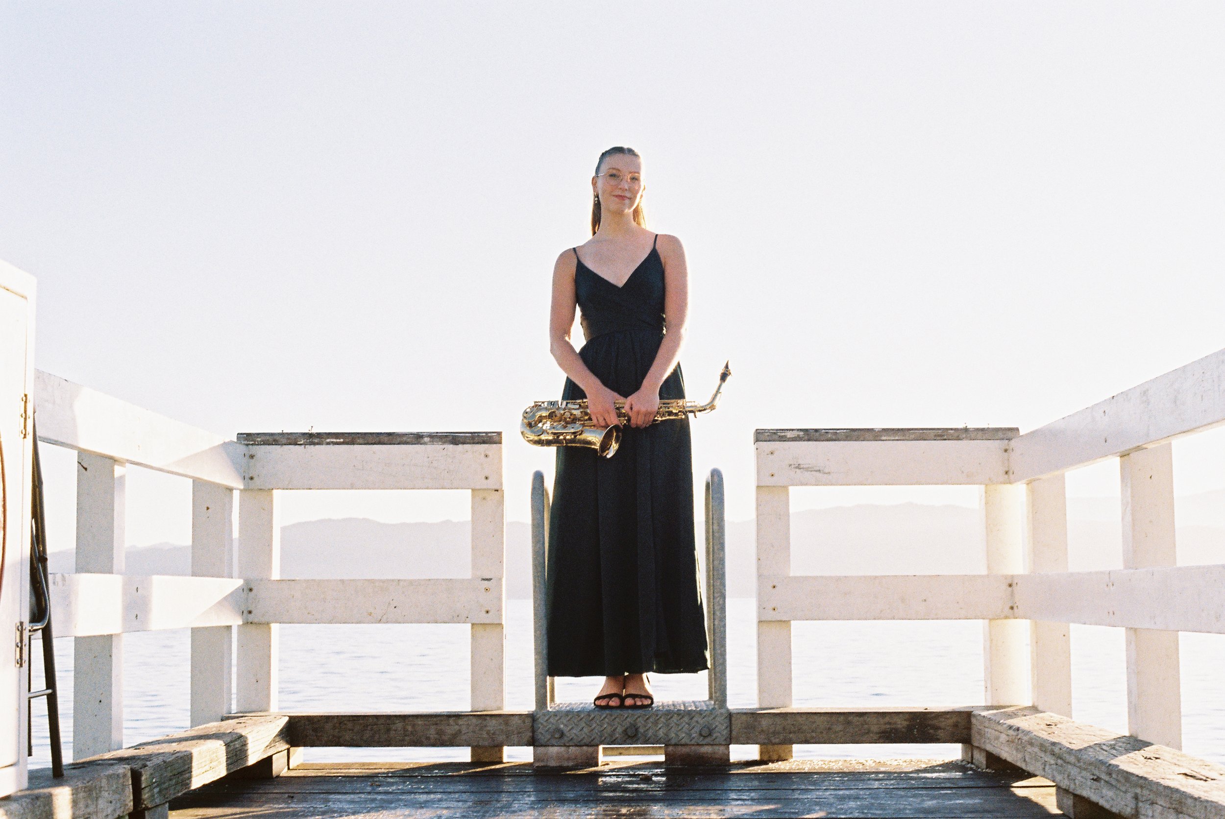 A woman in a black dress holding a saxophone standing on a dock by the water with white building structures in the background, under a bright sky.