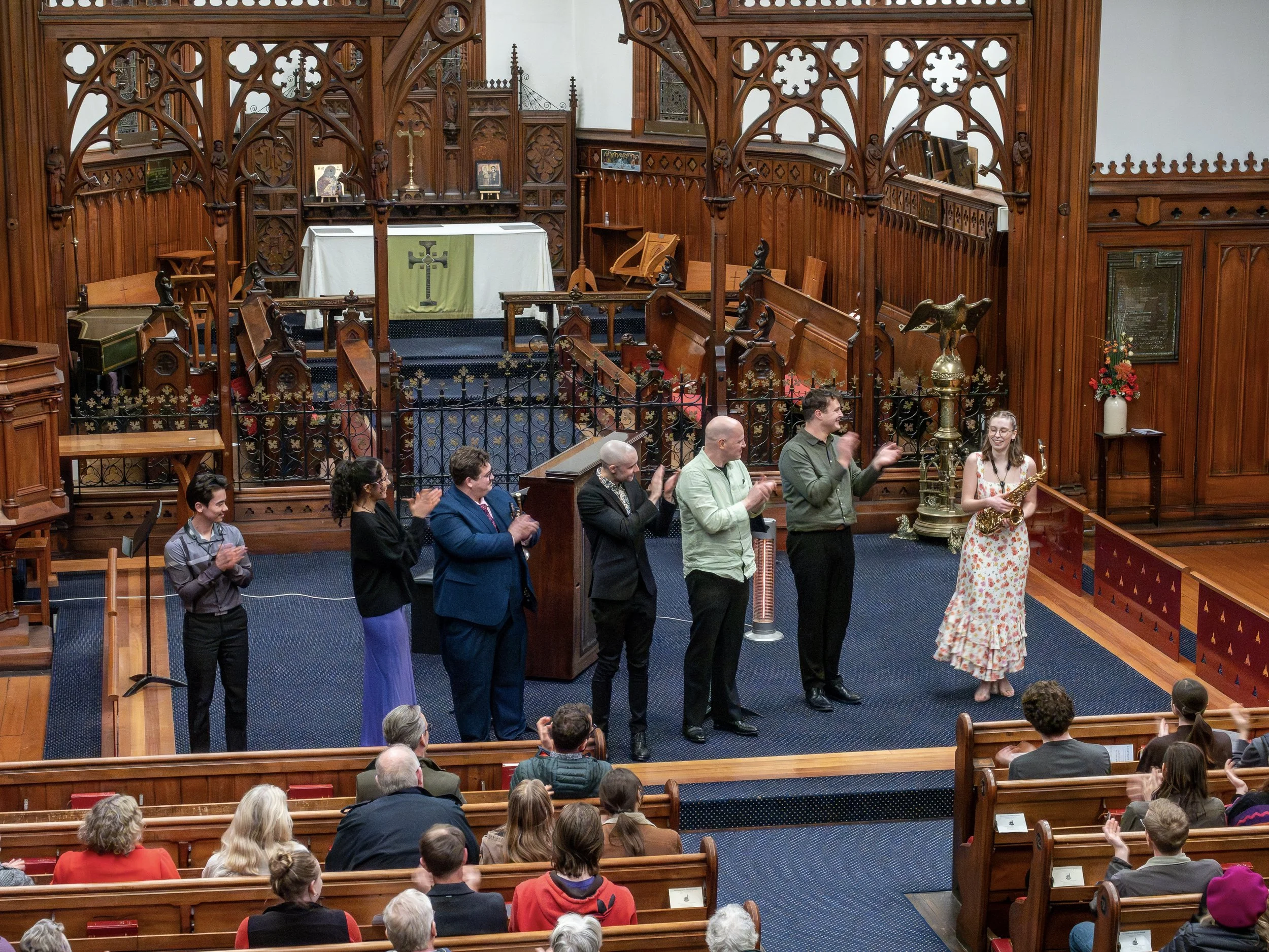 Group of people standing at the front of a church or chapel, some clapping, with a woman in a floral dress holding a saxophone. The interior features ornate wooden architecture and a religious altar.