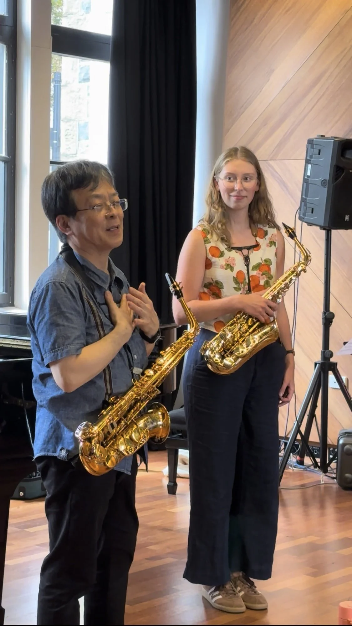 Two people holding saxophones during a musical performance or rehearsal, standing near a window and a wooden wall.