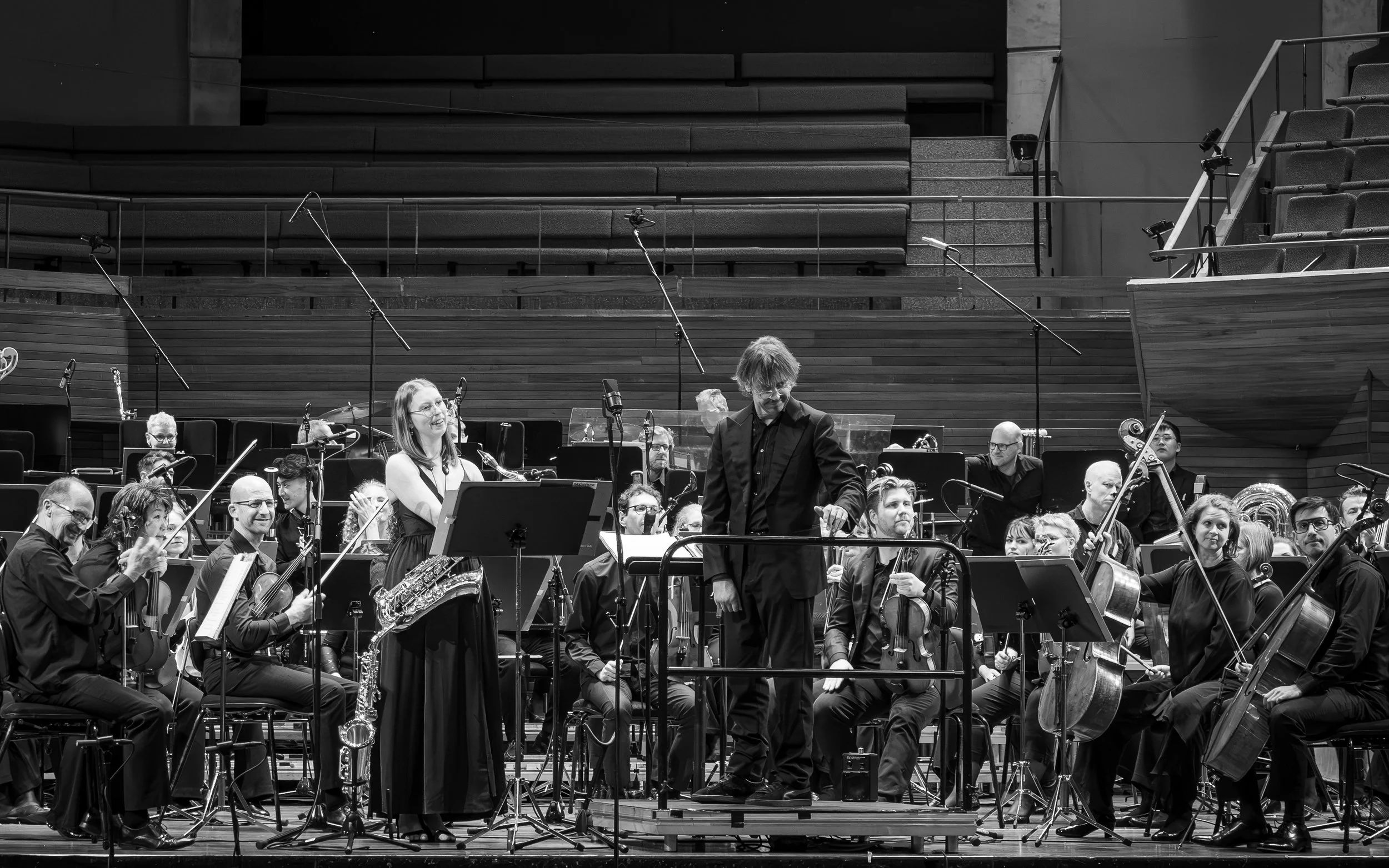 Black and white image of an orchestra performing on stage, with conductor interacting with musicians and various instruments visible.
