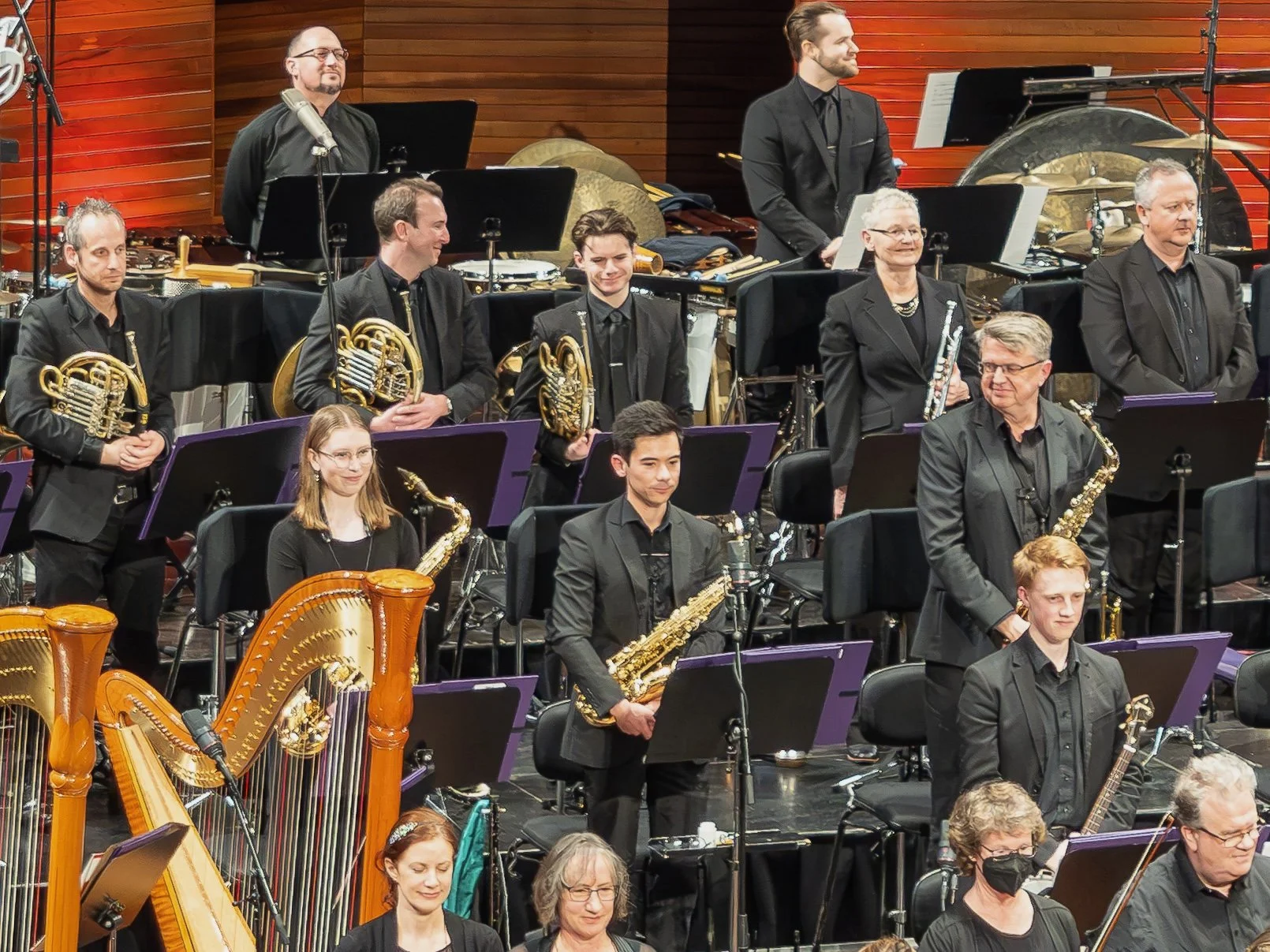 Orchestra members standing and seated on stage with musical instruments, wearing black formal attire, in a concert hall with wooden paneling.