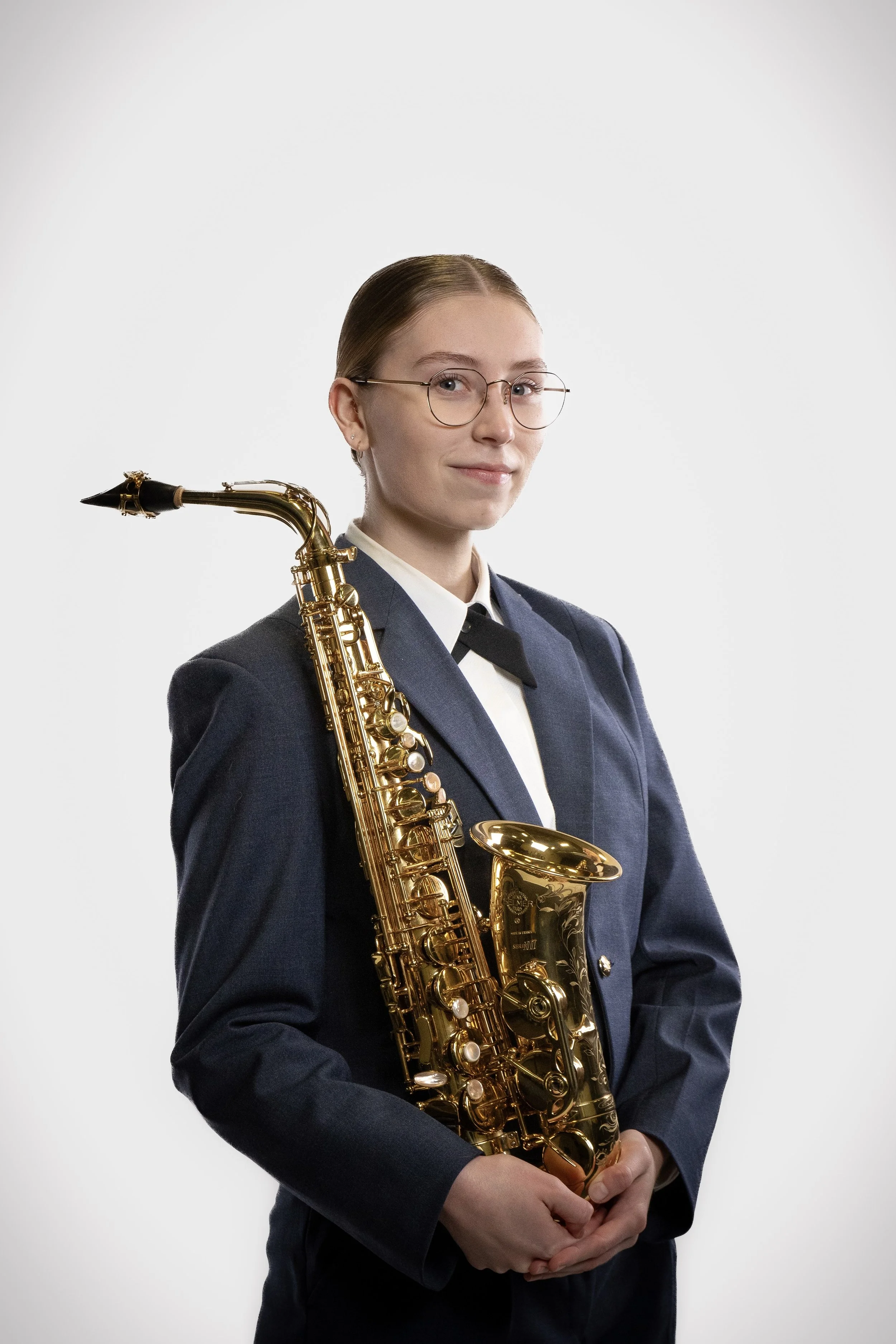 A woman wearing glasses and a dark blue blazer with a necktie, holding a golden saxophone against a light gray background.