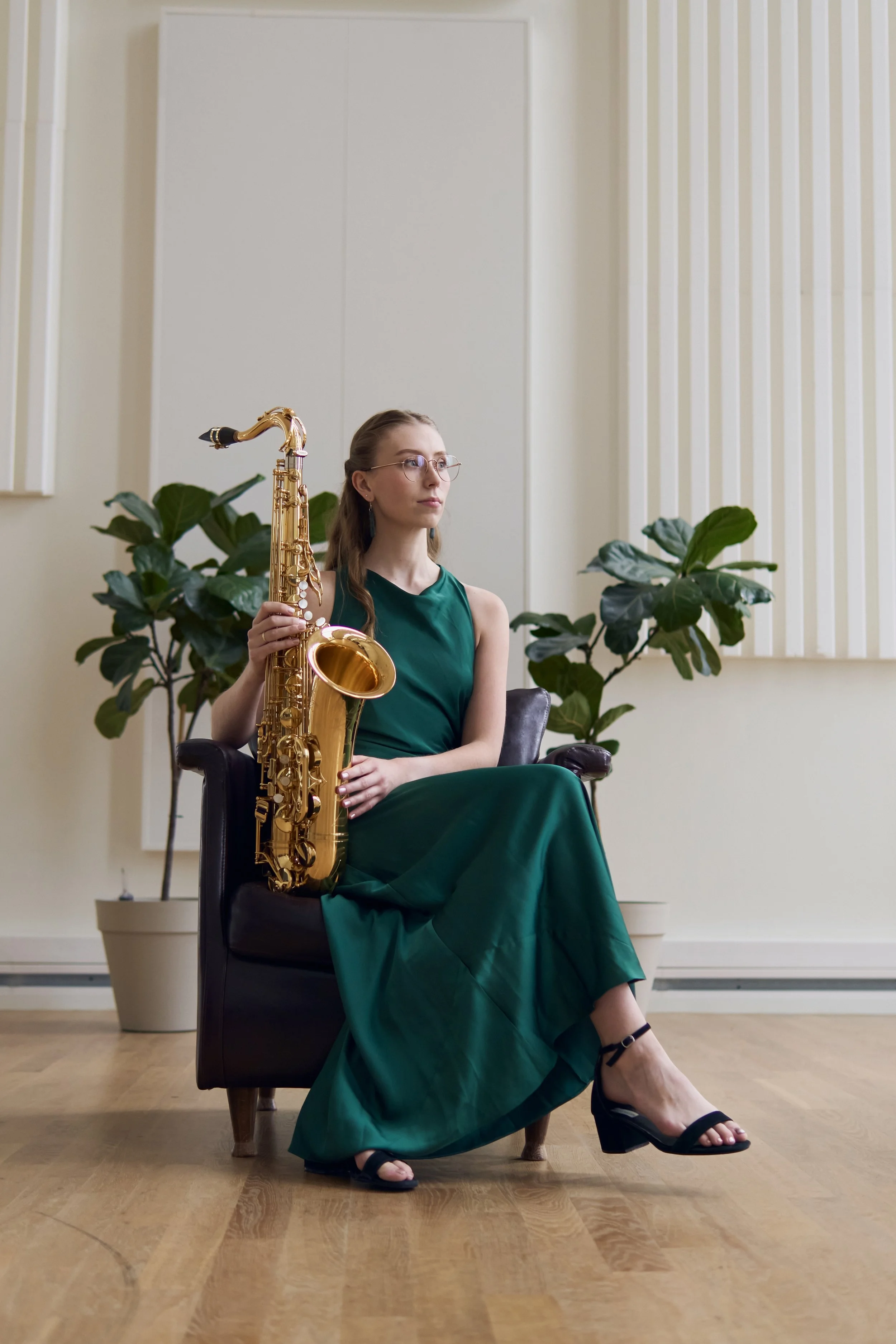 A woman in a long green dress with glasses holding a gold saxophone, sitting on a black chair in a room with wooden flooring and potted plants.