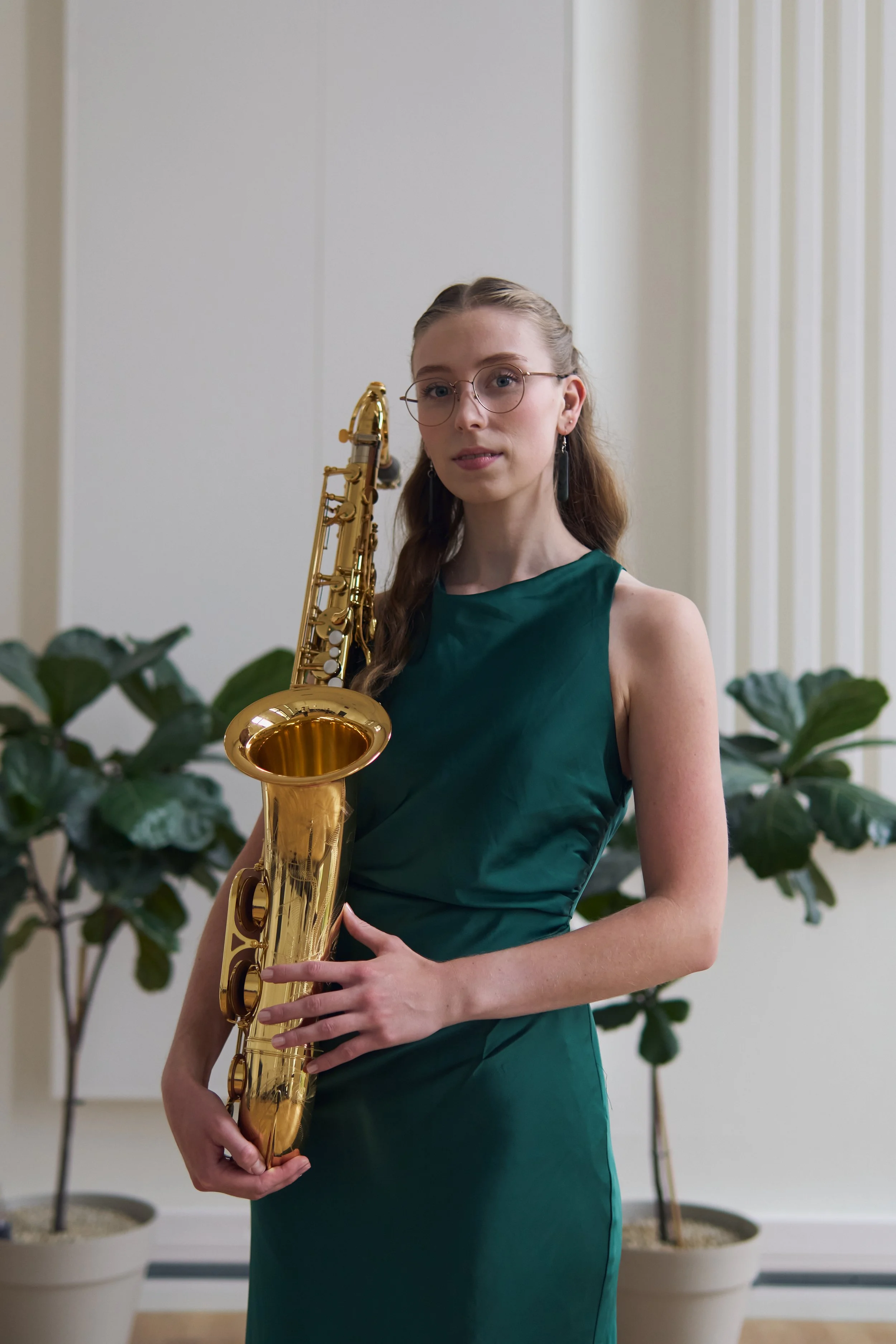 A woman wearing glasses in a teal dress holding a gold saxophone with potted plants in the background.