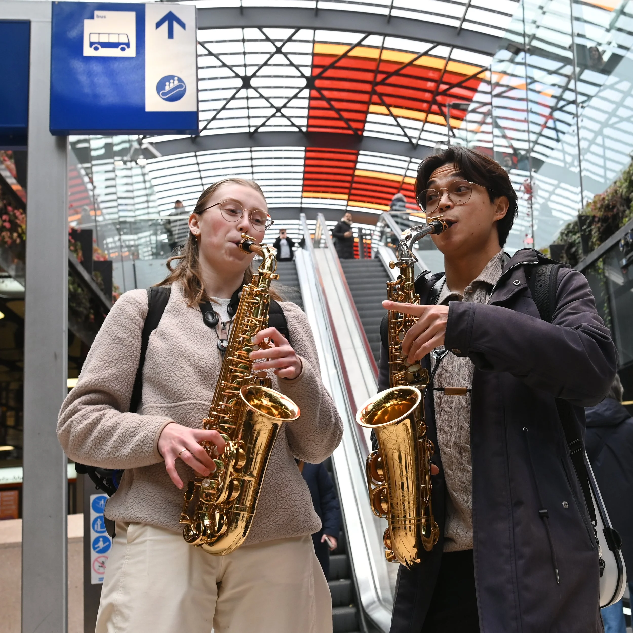 Two young adults are playing saxophones together in an indoor space, possibly a transit station, with a glass and metal ceiling overhead and a staircase with an escalator in the background.