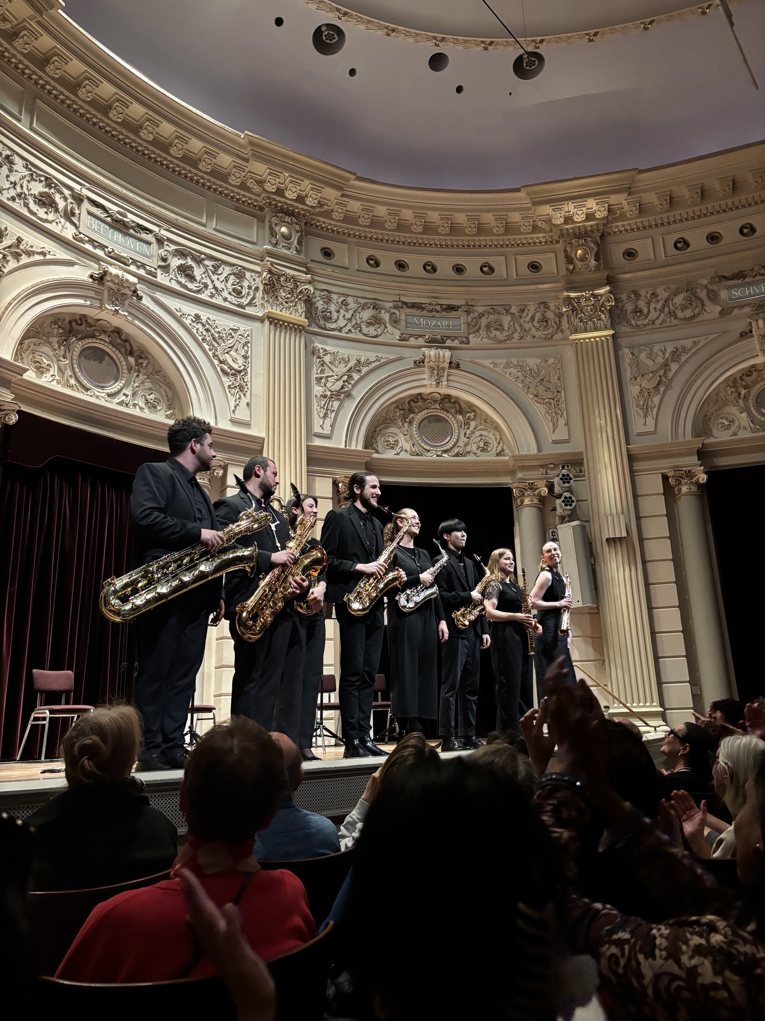 Orchestra standing on stage after a performance in an ornate concert hall, with audience members applauding.