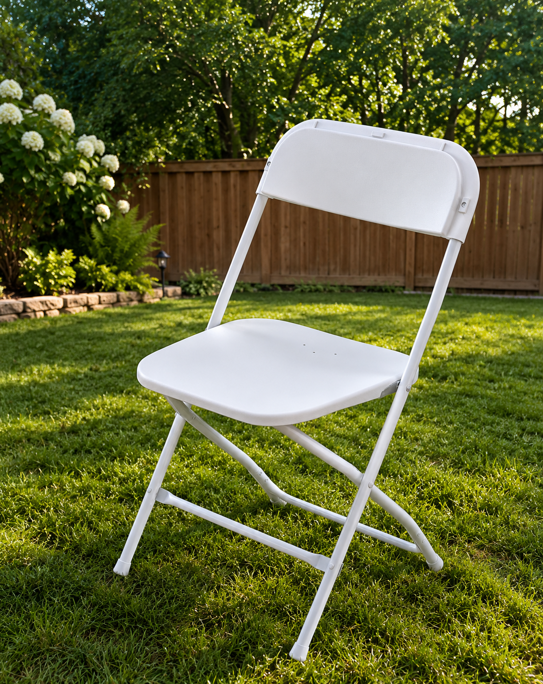 A white folding chair on a grassy backyard lawn with a wooden fence and green trees in the background.
