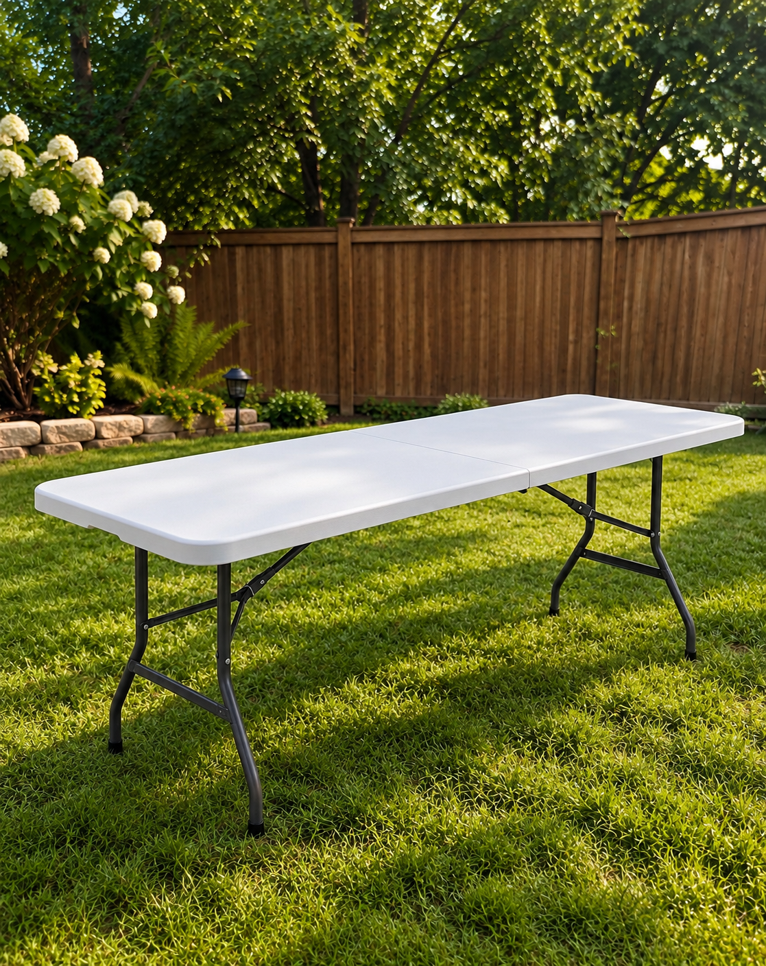 Empty white folding table set up on green grass in a backyard with a wooden fence, trees, and flowering bushes in the background.