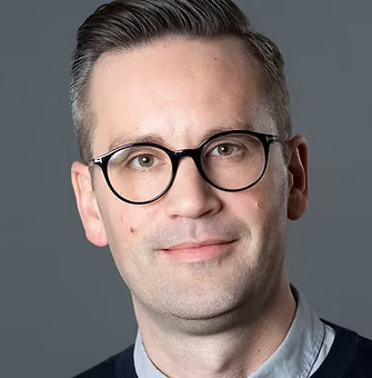 Porträtt av en man med glasögon, ljus hud, mörkt hår och kläder i mörka och ljusa nyanser.