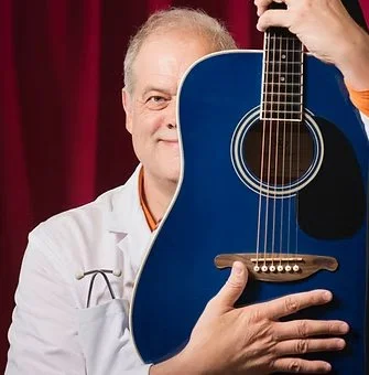 En man i vit läkarkläder håller en blå akustisk gitarr framför ansiktet, med en röd sammetshimmel i bakgrunden.