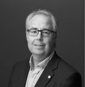 Svartvitt porträtt av man med glasögon, grått hår, blå skjorta och mörk kostym.