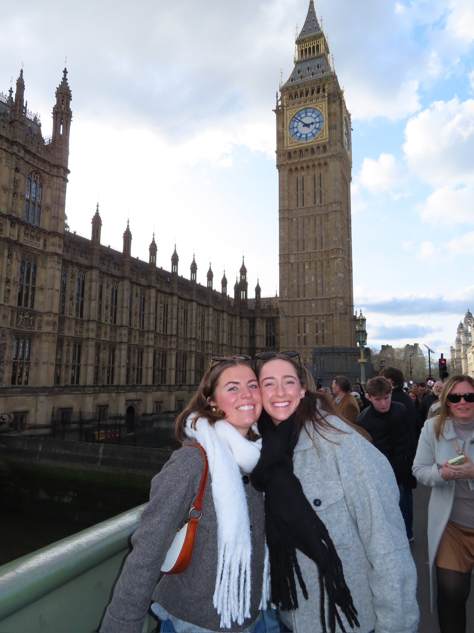 Two young women stand close together on a bridge in front of the Big Ben clock tower in London, smiling at the camera. Other people are visible in the background.