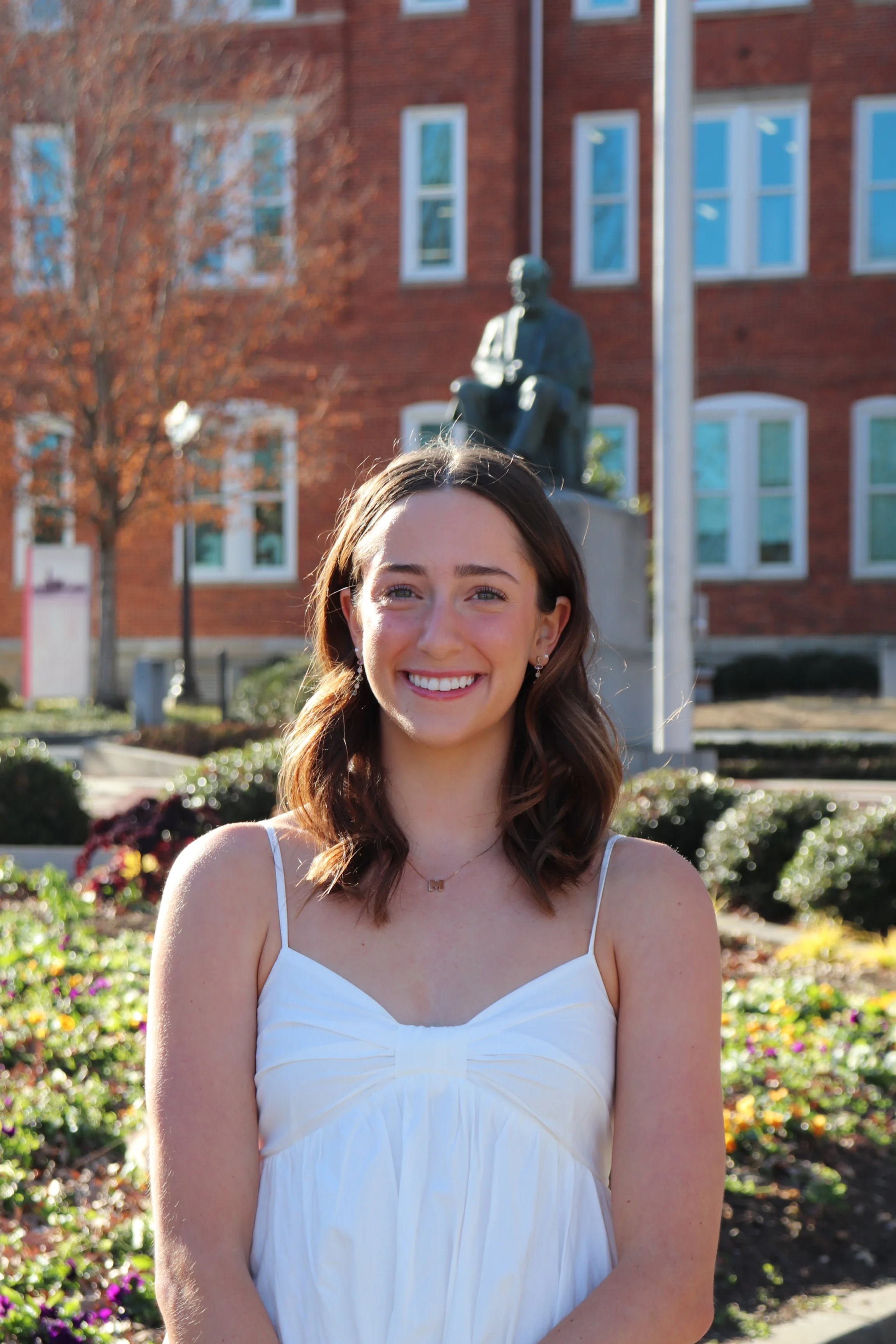 A smiling young woman with shoulder-length brown hair, wearing a white dress with spaghetti straps, standing outdoors in front of a garden and a statue of a seated man. There is a brick building with multiple windows and some trees in the background.