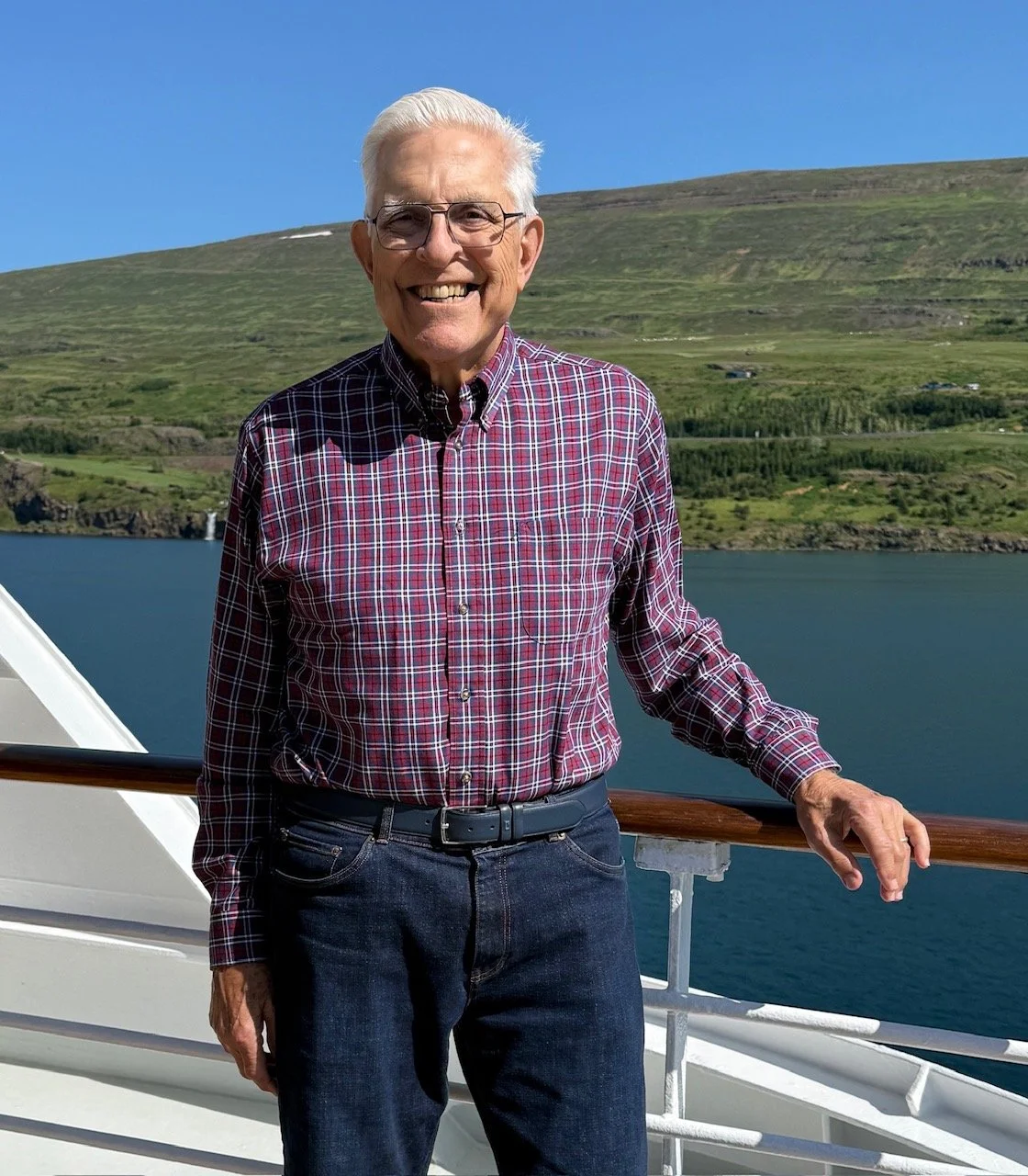 A man with glasses and white hair smiling on the deck of a boat with water, green hills, and a clear blue sky in the background.
