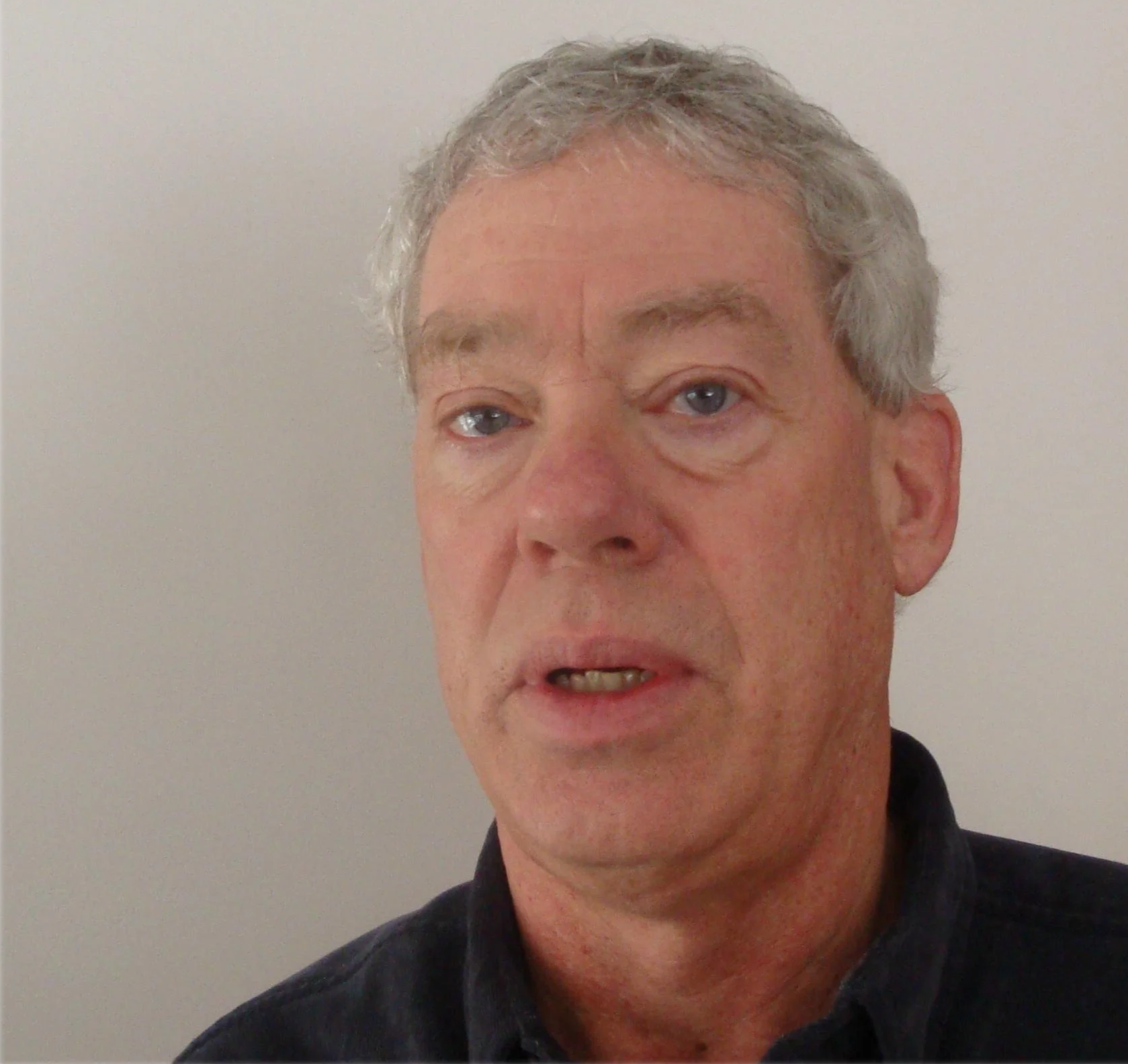 A man with gray hair and blue eyes, speaking or making a facial expression, wearing a dark collared shirt against a plain white wall background.