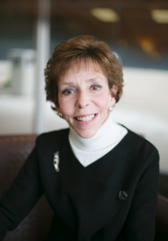 A woman with short brown hair, wearing a black blazer and white turtleneck, smiling while sitting indoors.