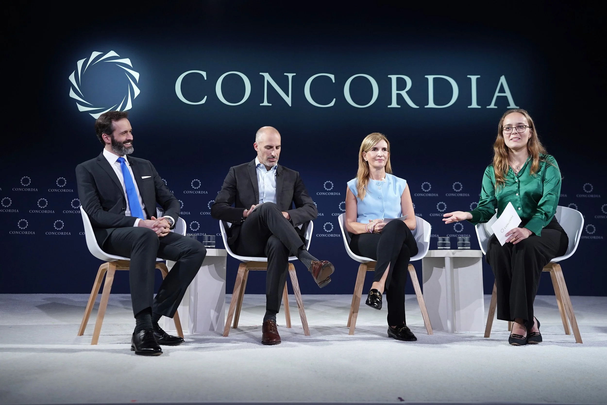 Four people sitting on chairs and engaging in a panel discussion or interview on stage with a large blue backdrop displaying the word 'CONCORDIA' and a circular logo.