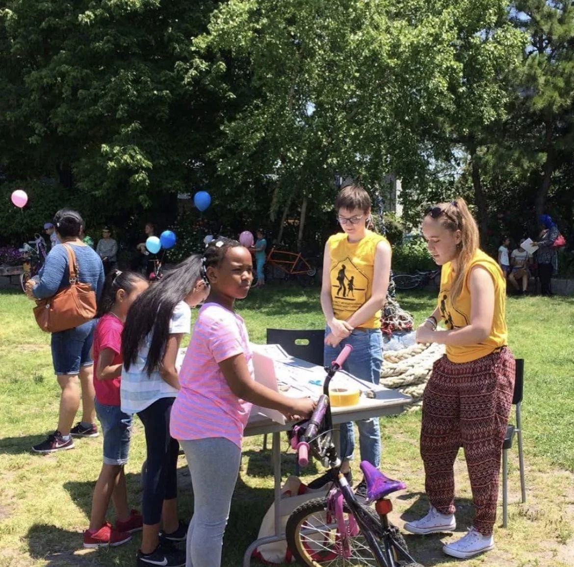 Children and adults gathered outdoors under trees around a table with items, some holding balloons, in a park-like setting during a community event.