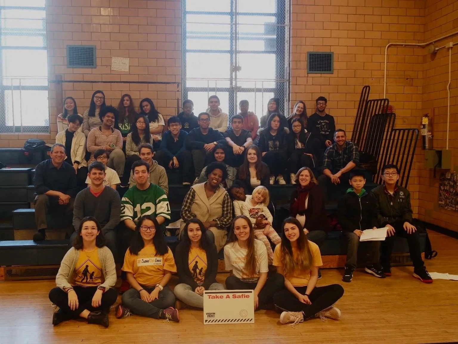 Group of diverse people posing in a gymnasium with a sign that reads 'Take A Safie' in the front.