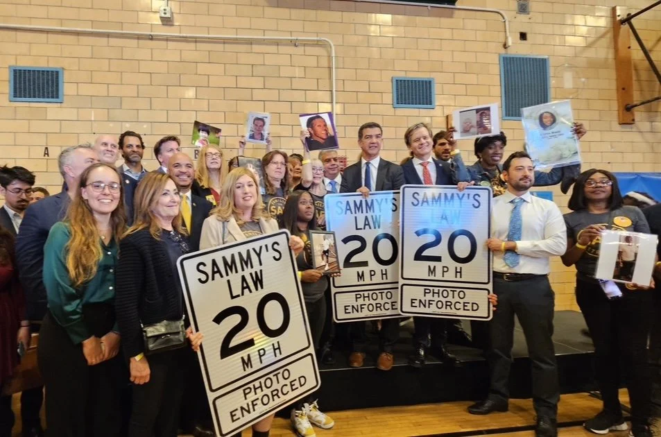 Group of people holding signs that read "SAMMY'S LAW 20 MPH PHOTO ENFORCED," at an indoor event, with some holding framed photos of individuals, in front of a brick wall.
