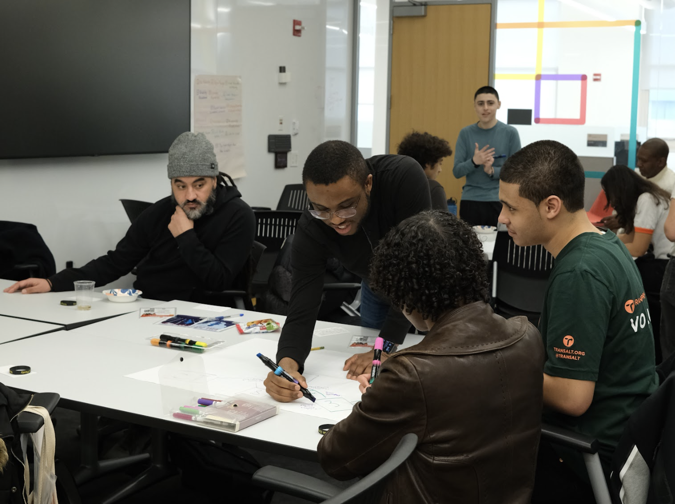 Group of diverse young adults working together around a table in a classroom or conference room, engaging in a collaborative activity with markers and papers, with a person standing at the back speaking.
